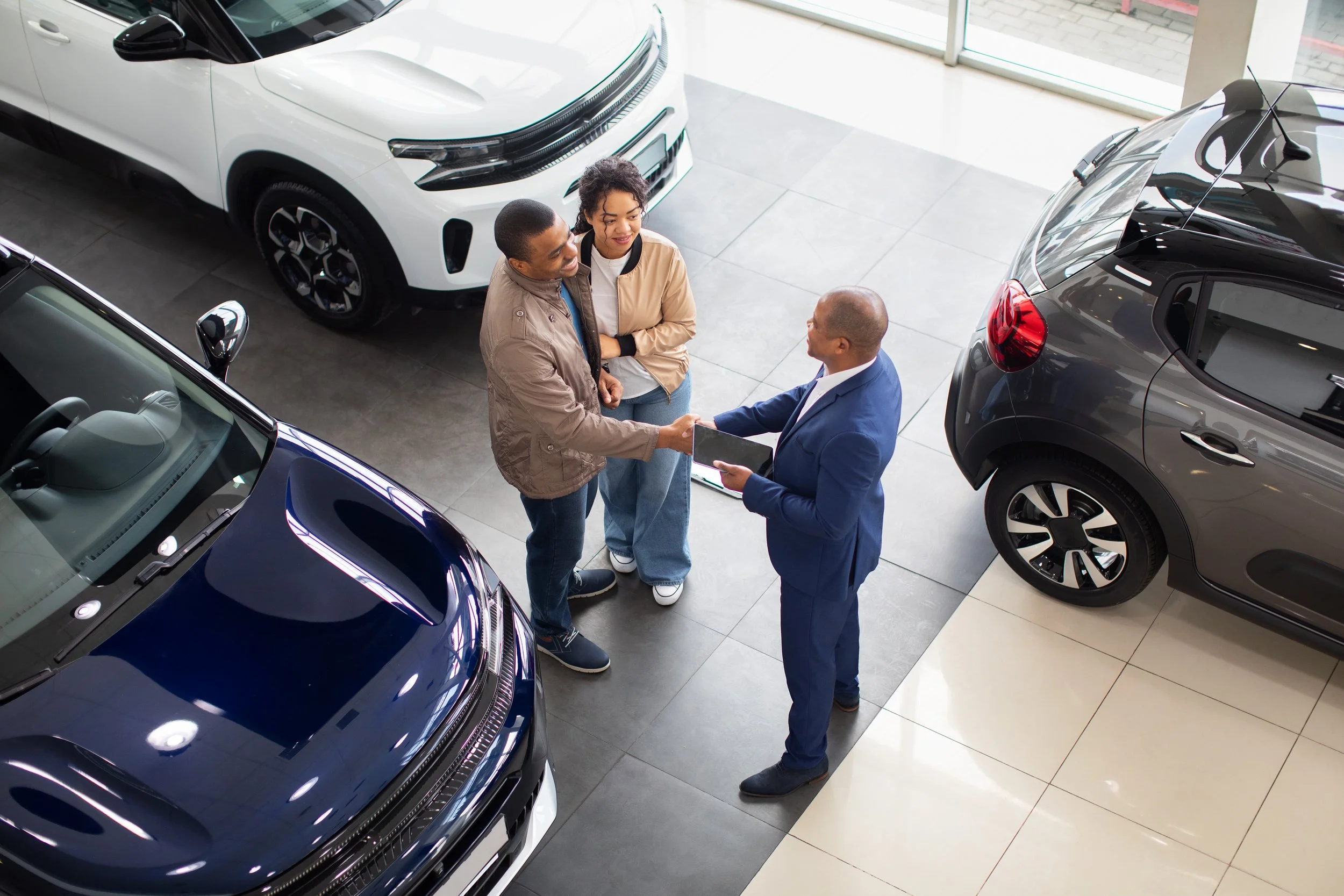 A man and woman shaking hands with a car sales representative inside a car dealership, with multiple cars around them.