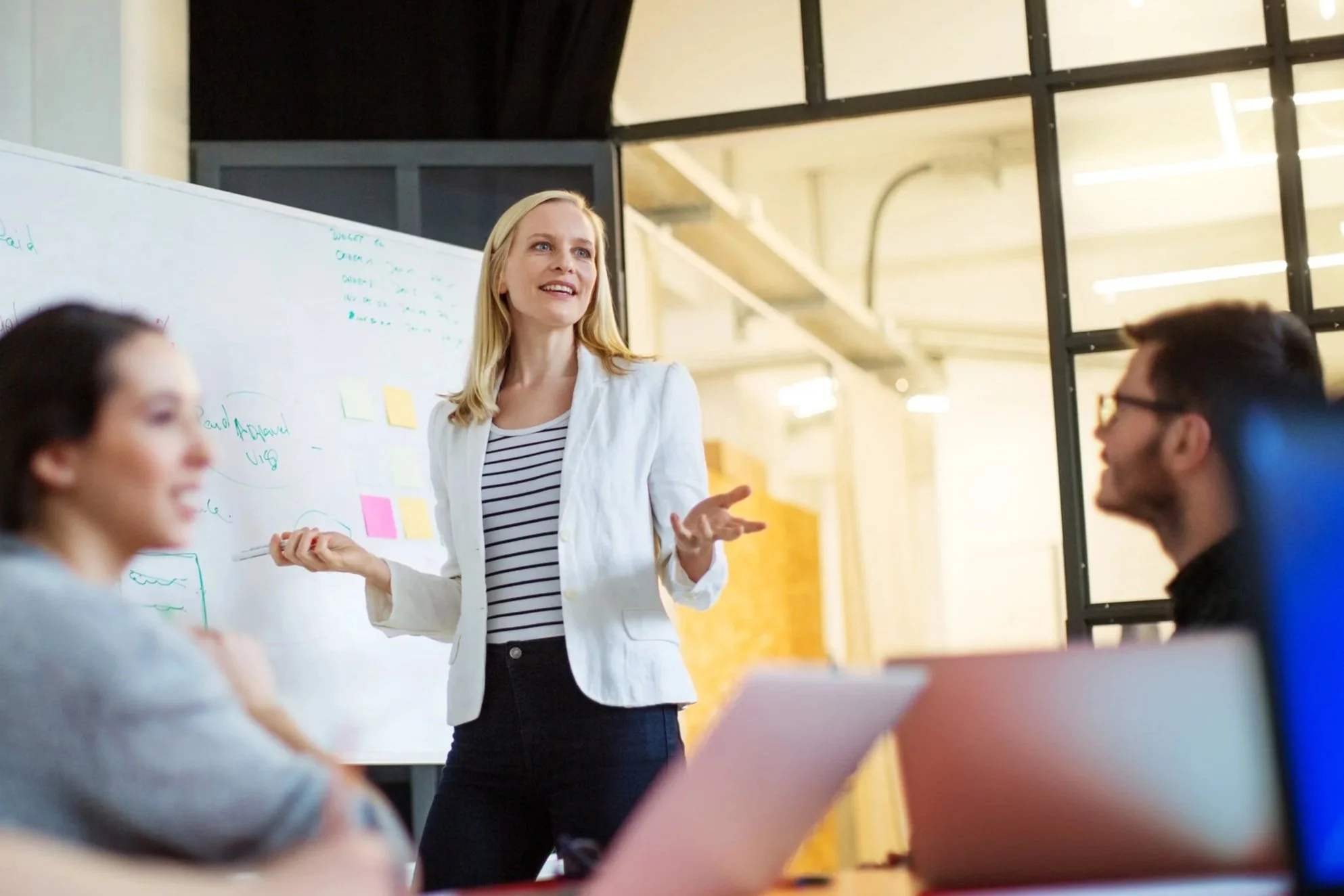 A woman standing in front of a whiteboard with writing and colorful sticky notes, speaking to a group in a modern office or classroom setting.