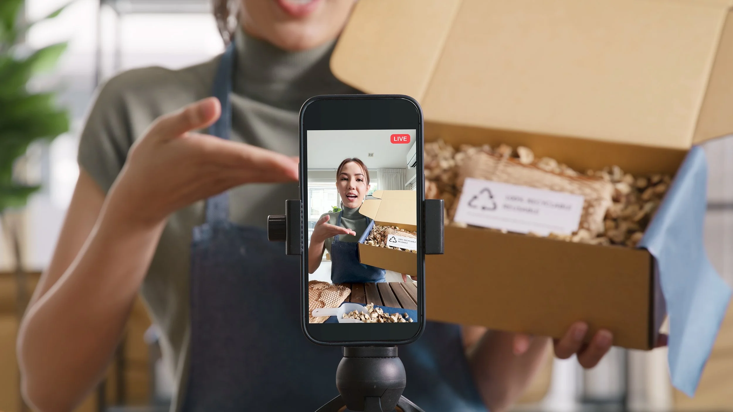 A person is recording a live video on a smartphone, which shows a woman with brown hair in a gray turtleneck sleeve, gesturing towards a box of recyclable packing material on a wooden table.