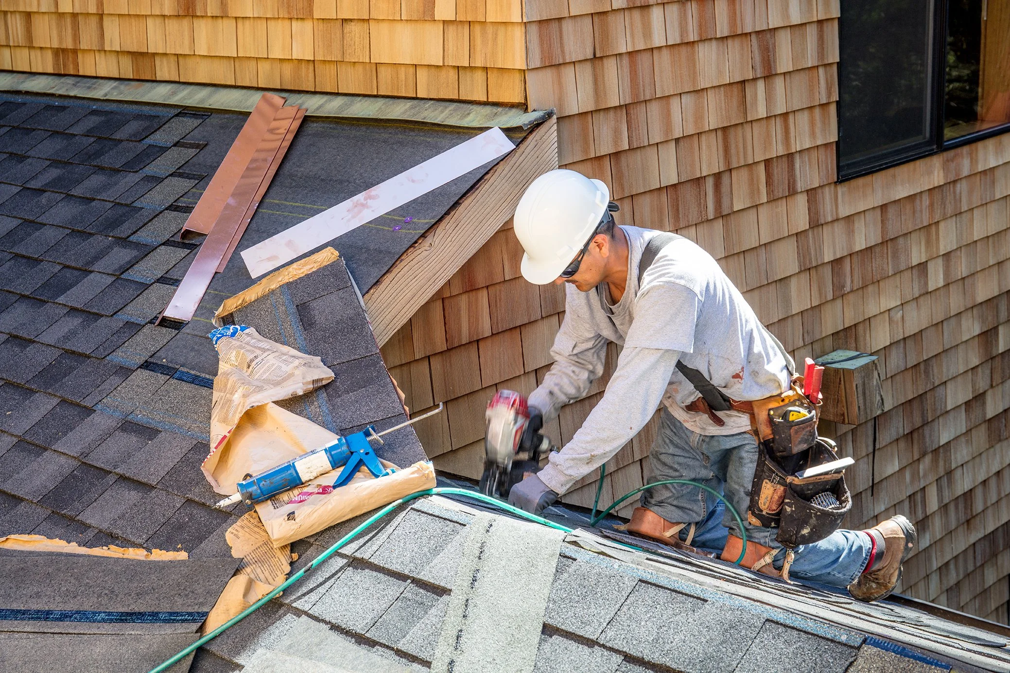 A construction worker wearing a white hard hat and sunglasses is working on a roof, installing or repairing shingles. There are tools, a caulking gun, and materials around him, with a wooden house's exterior wall in the background.