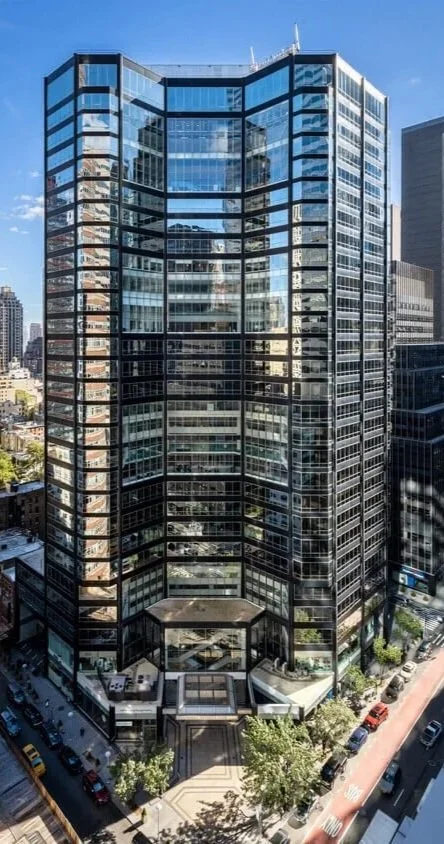 Tall modern glass office building in an urban cityscape, reflecting surrounding skyscrapers and clear blue sky.