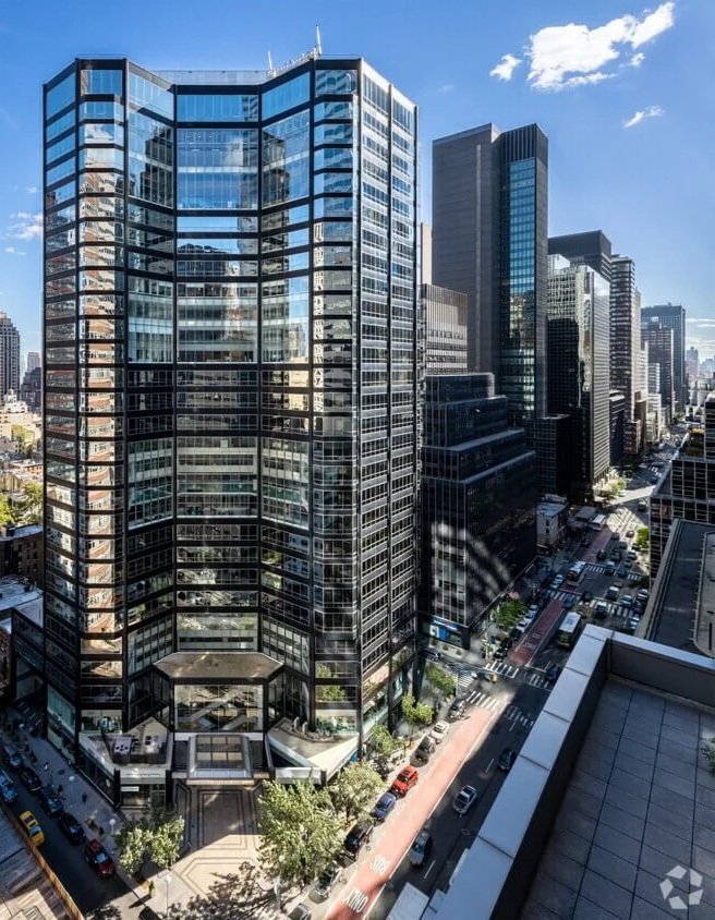 A tall modern glass office building in a city with other skyscrapers, a busy street below with cars and trees, under a partly cloudy sky.