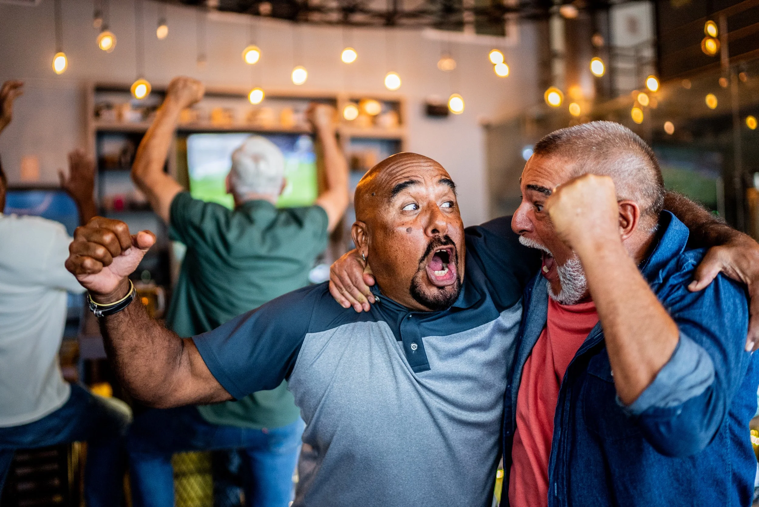 Two men are celebrating and cheering at a social gathering. One man, with a shaved head and a goatee, is wearing a gray and black shirt, and the other man has long hair tied back and is wearing a blue jacket with a red shirt underneath. They are embracing and showing excitement. In the background, there are other people and a television screen.