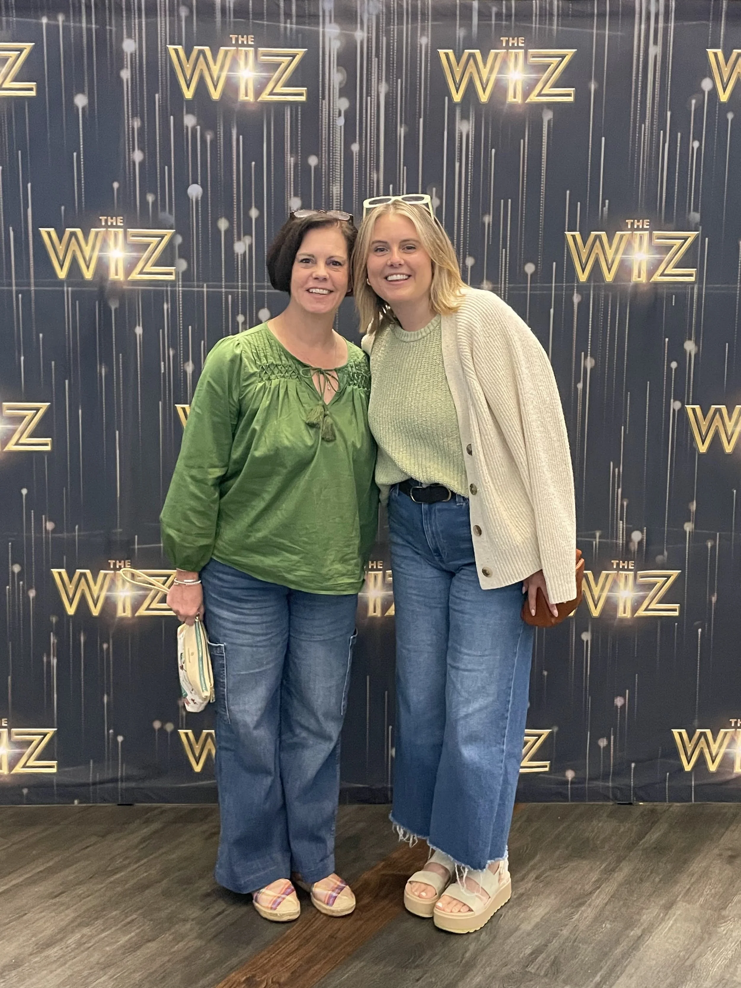 Two women smiling and posing together in front of a backdrop with the text 'The Wiz' repeated across it. They are dressed casually, one in a green top and the other in a light-colored cardigan, both wearing jeans and platform shoes.