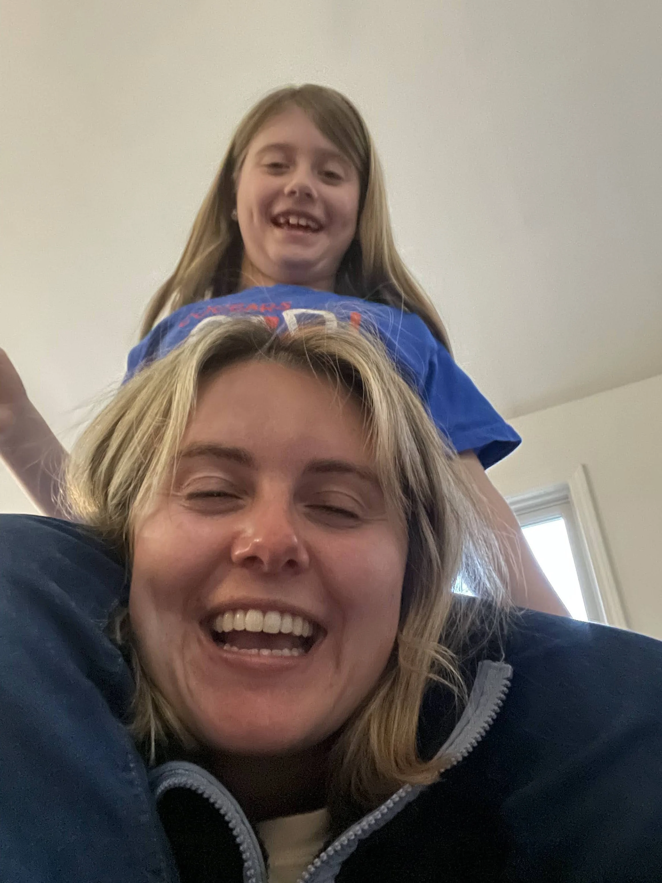 A woman with blonde hair smiling widely, lying down with a child on her back. The child, with light brown hair, is smiling and leaning forward. The background shows a white wall and a window.
