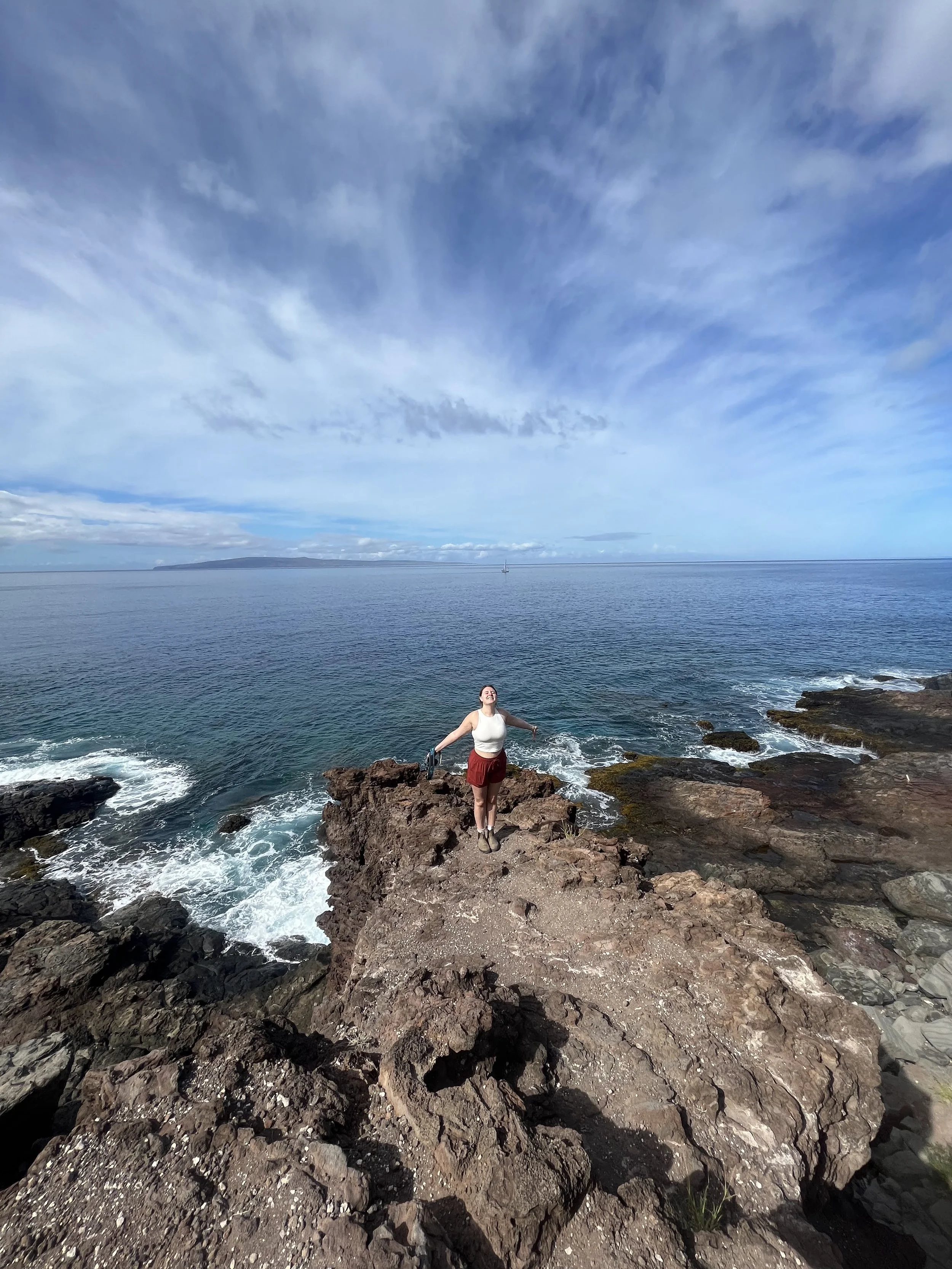 R Photos photographer Rachel Kosky standing on volcanic coastal rocks with arms wide open, ocean stretching to the horizon — adventure-loving wedding and elopement photographer available for destination shoots
