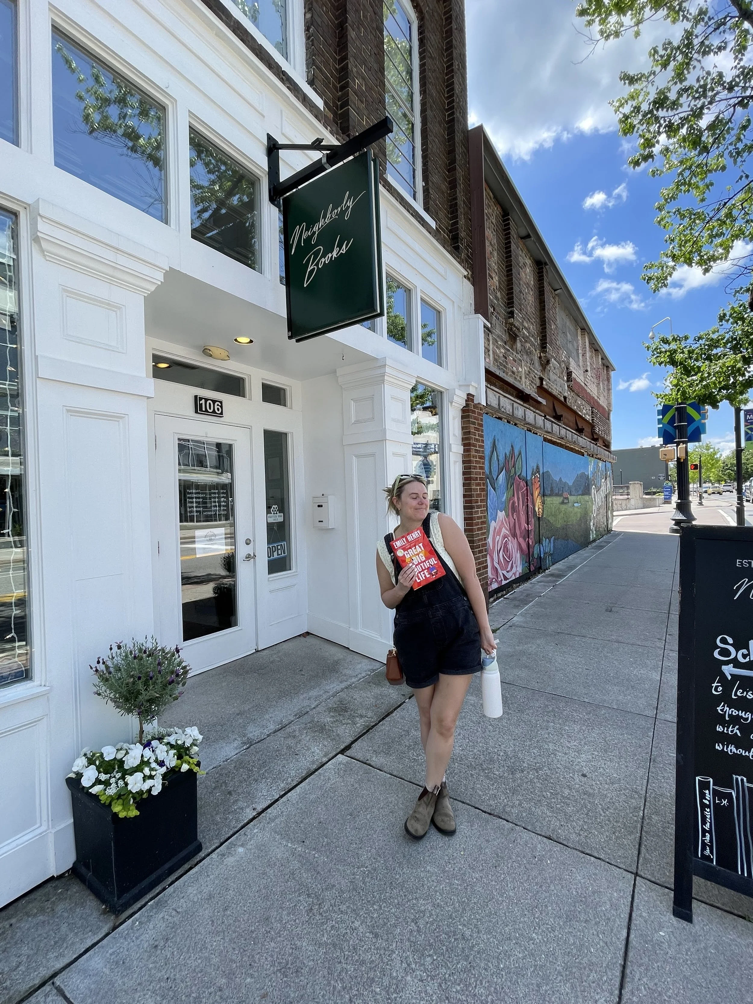 Rachel Kosky, Tennessee photographer, walking outside Neighborly Books with a book and water bottle on a sunny day — personal brand photo for R Photos in Maryville TN