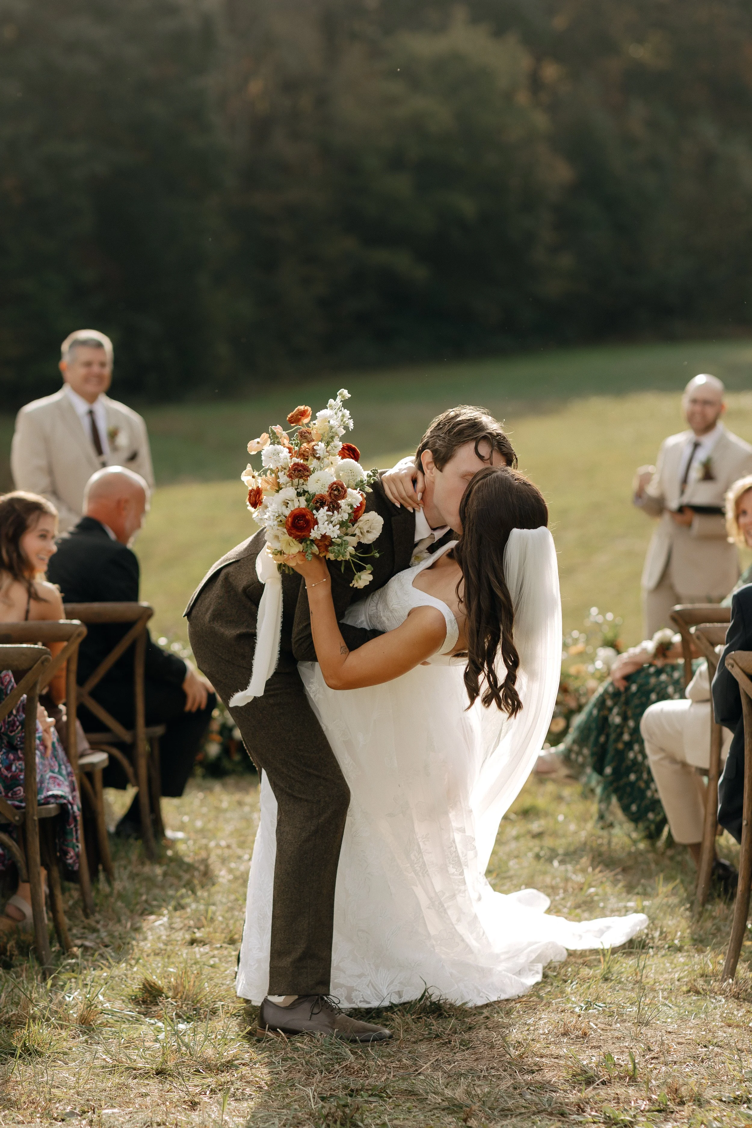 Groom dipping bride for a first kiss at an outdoor wedding ceremony in North Georgia, surrounded by guests seated in wooden chairs, with a lush green meadow and forest backdrop — Tennessee wedding photographer