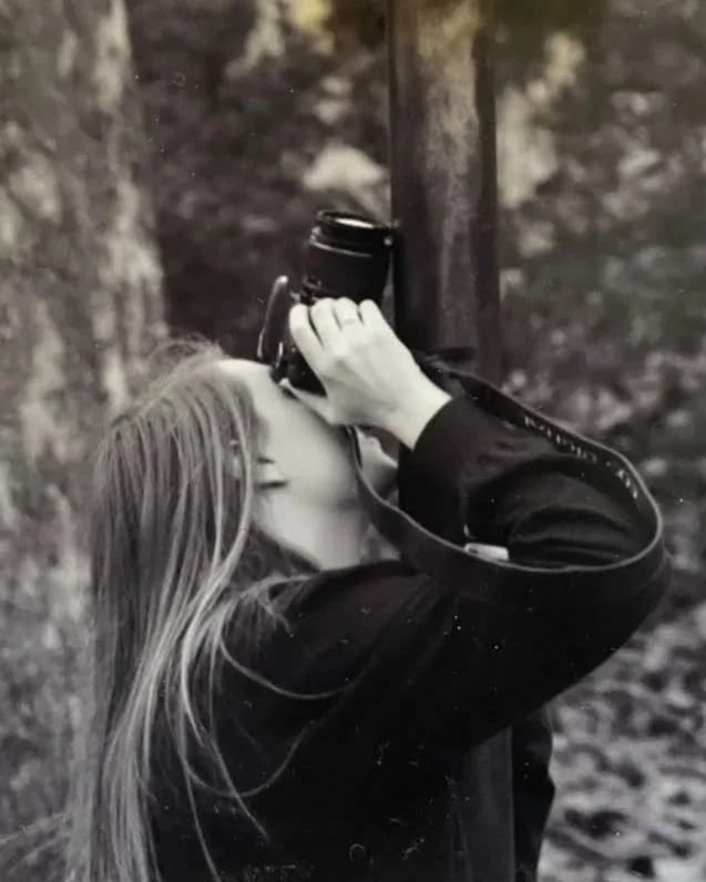 A woman with long hair is taking a photograph outdoors with a camera, standing near a tree trunk with a blurry natural background.