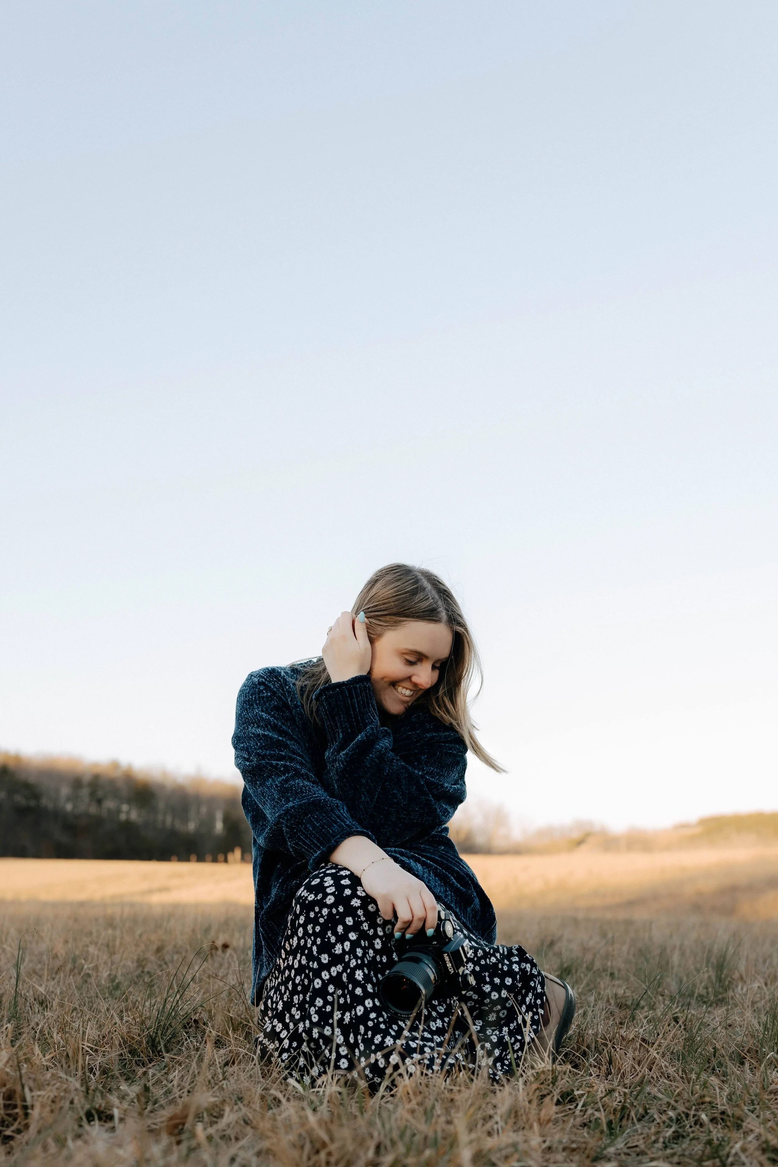 Candid portrait of R Photos photographer Rachel Kosky seated in a dry winter meadow at golden hour, smiling down at her camera — documentary wedding and elopement photographer in Chattanooga, TN