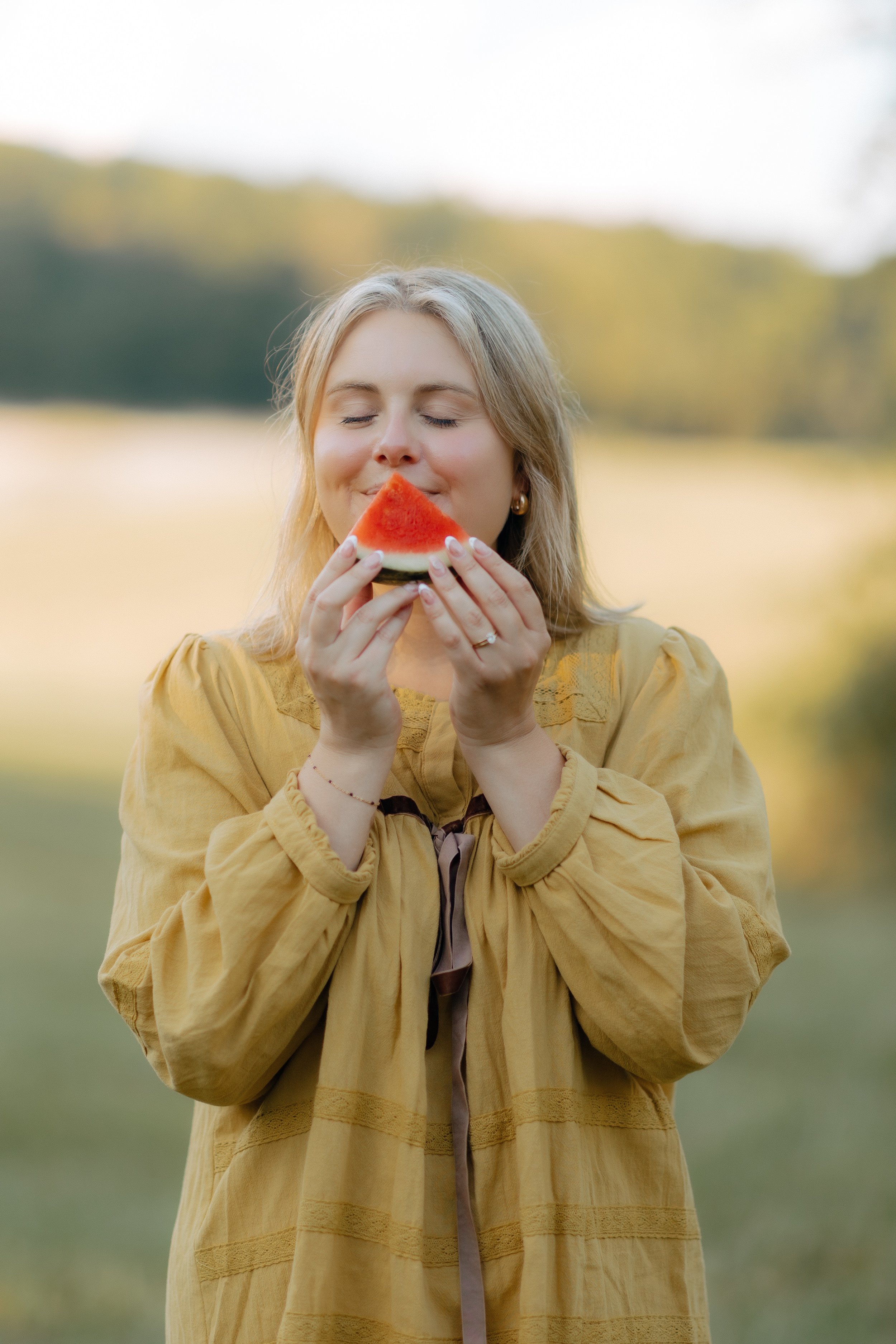 Warm golden hour portrait of R Photos photographer Rachel Kosky in a mustard dress enjoying a watermelon slice in a Tennessee meadow — approachable, joyful wedding photographer based in Chattanooga, TN