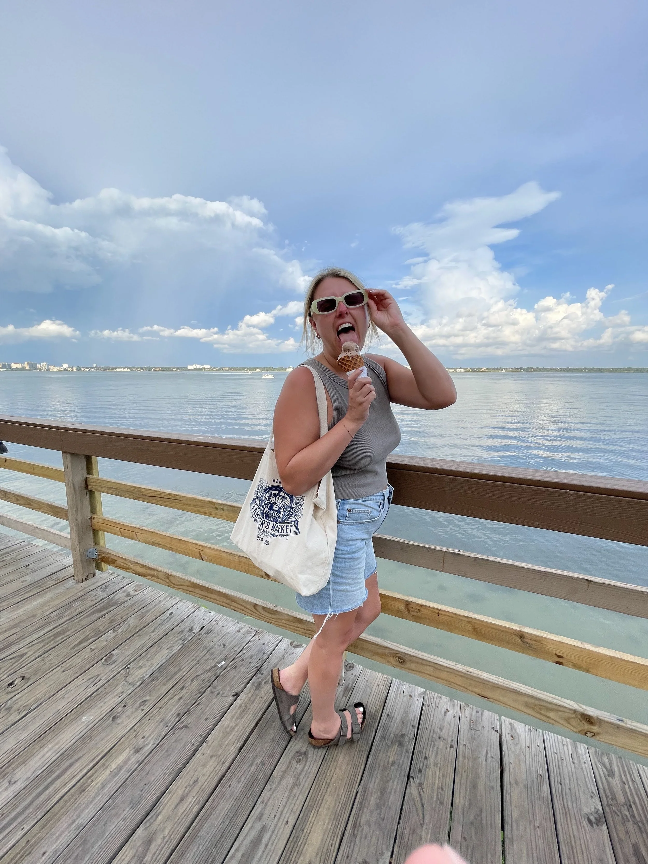 Candid and playful photo of R Photos photographer Rachel Kosky enjoying an ice cream cone on a sunny pier, tote bag over her shoulder and sunglasses in hand — the real person behind the camera