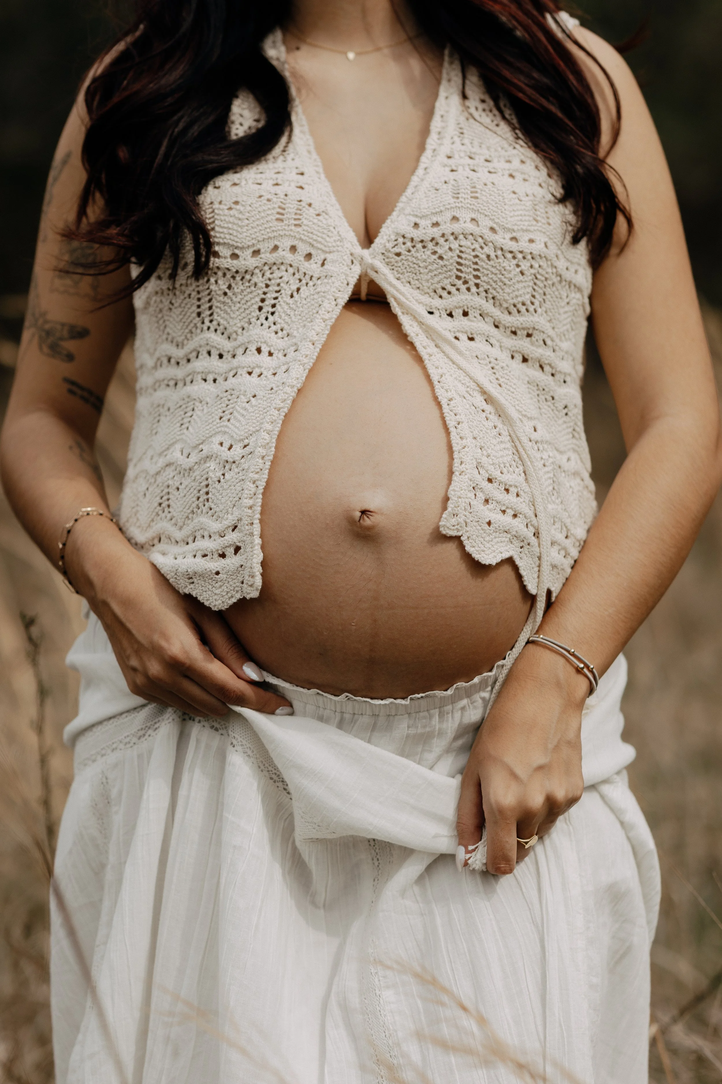 Boho Pregnant woman showing belly during golden hour in a field of tall grass wearing crochet top - Boho Maternity Photographer