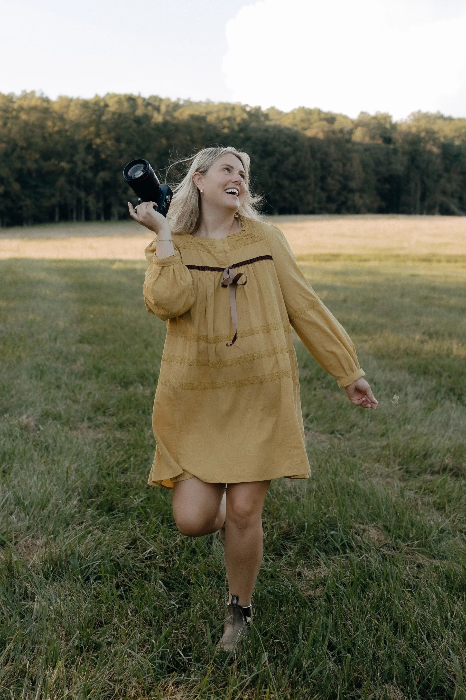 Smiling photographer in a yellow dress holding a camera in a golden hour field, Chattanooga TN photography studio R Photos