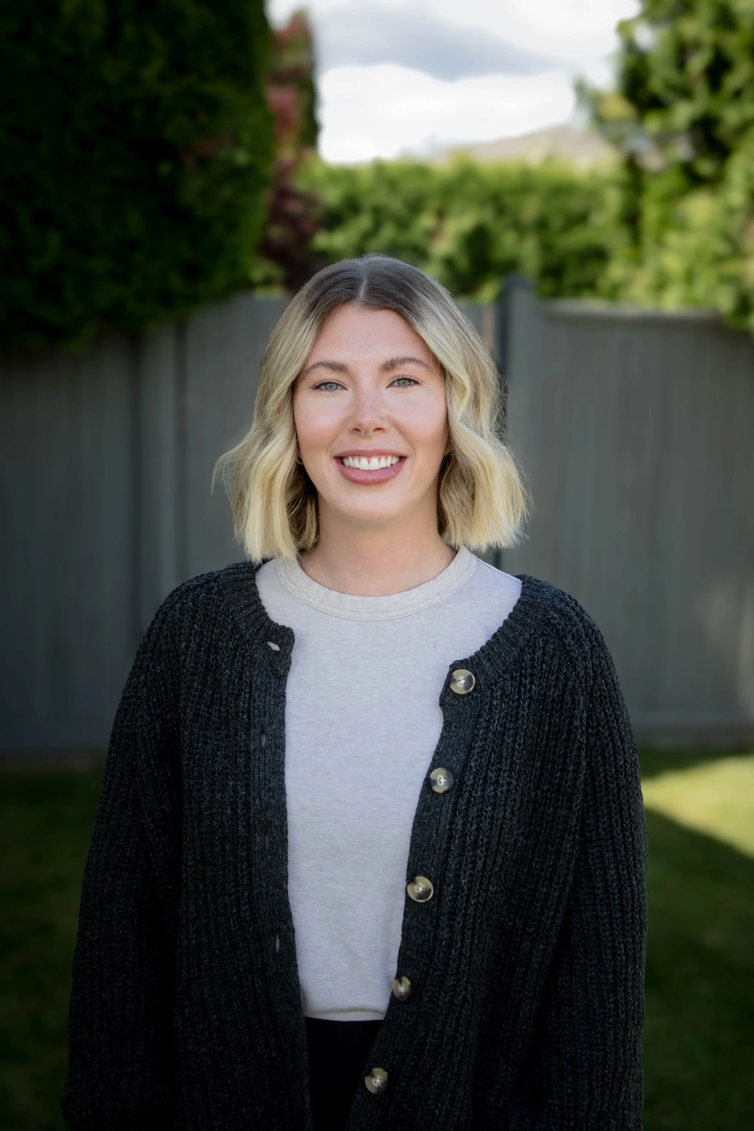 A young woman with shoulder-length blonde hair and a nose ring, smiling outdoors with a blurred green hedge and sky in the background.
