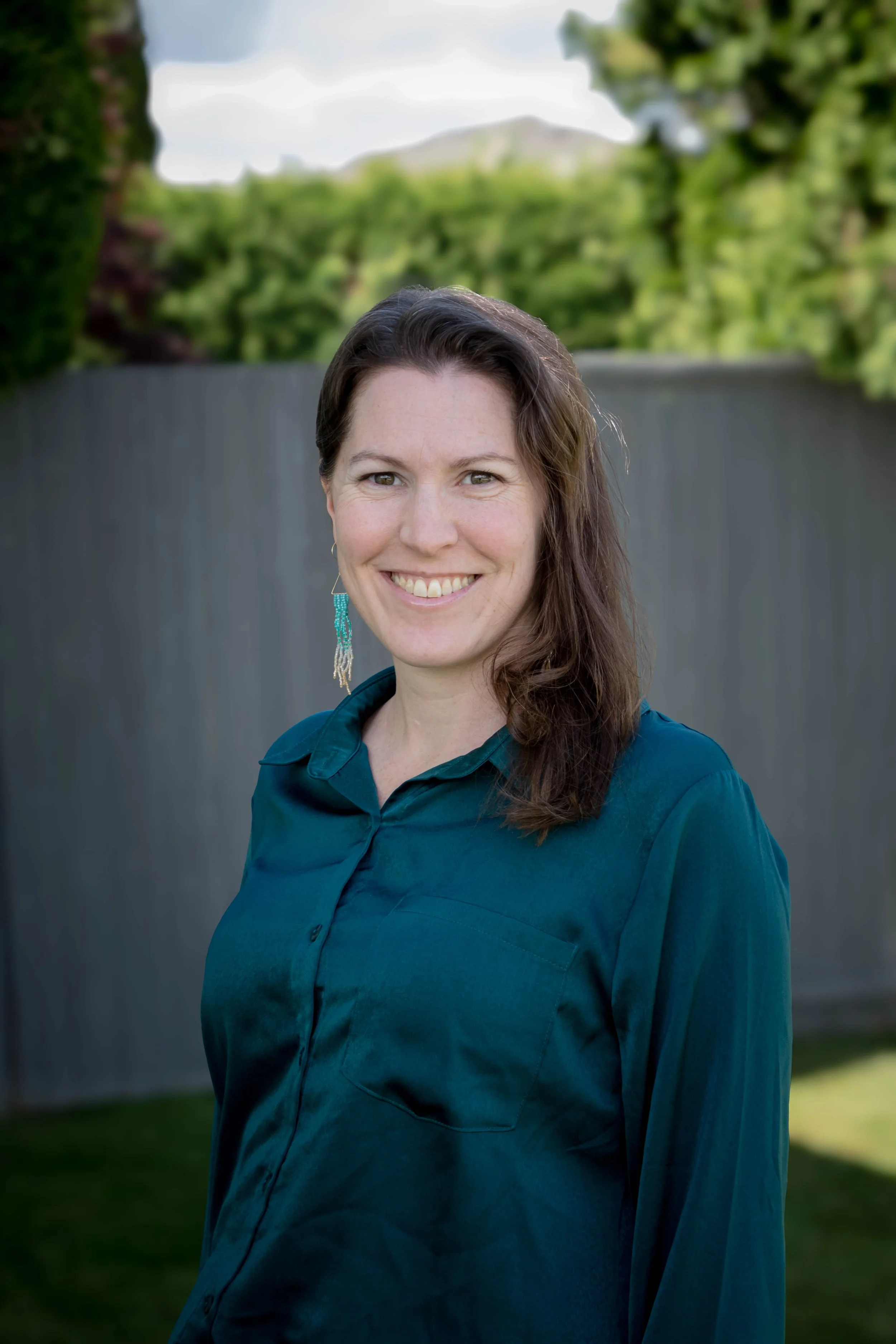 A woman with shoulder-length brown hair, smiling, wearing a dark teal button-up shirt with a collar, standing outdoors in front of a gray fence and green trees with a blurred mountain in the background.