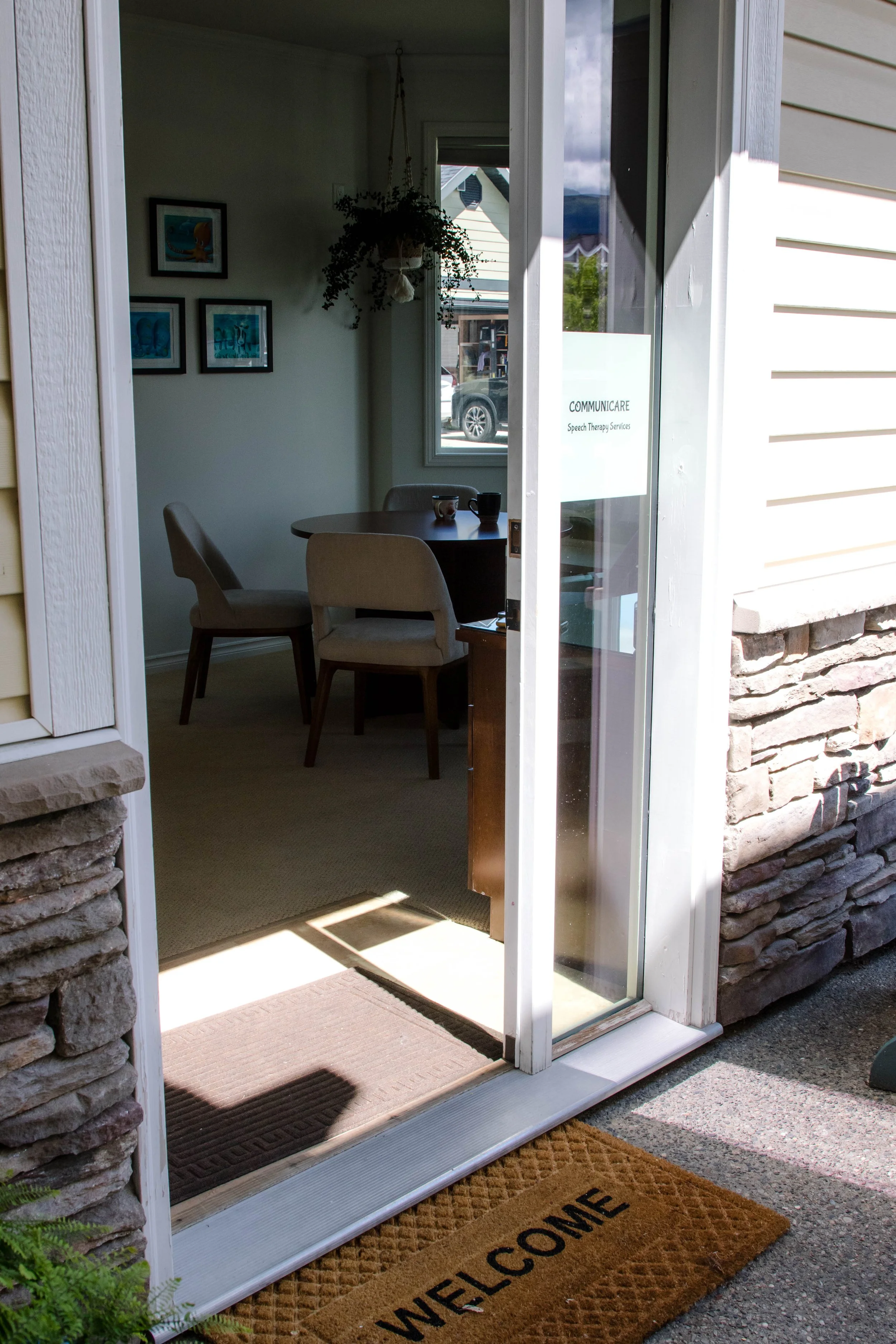 Looking into a cozy therapy area through a glass door, with a round table, three beige upholstered chairs, and framed artwork on the wall. A welcome mat is at the door entrance.