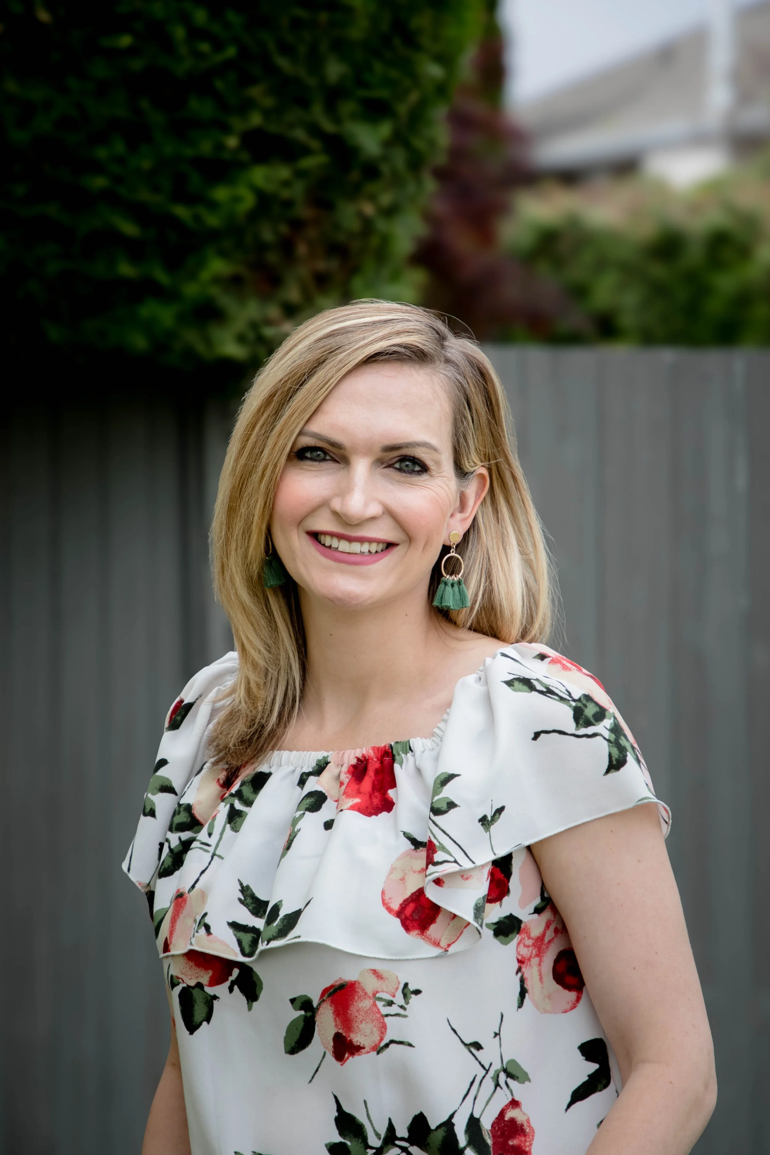 A woman with blonde hair and earrings smiling outdoors in front of a wooden fence and greenery.