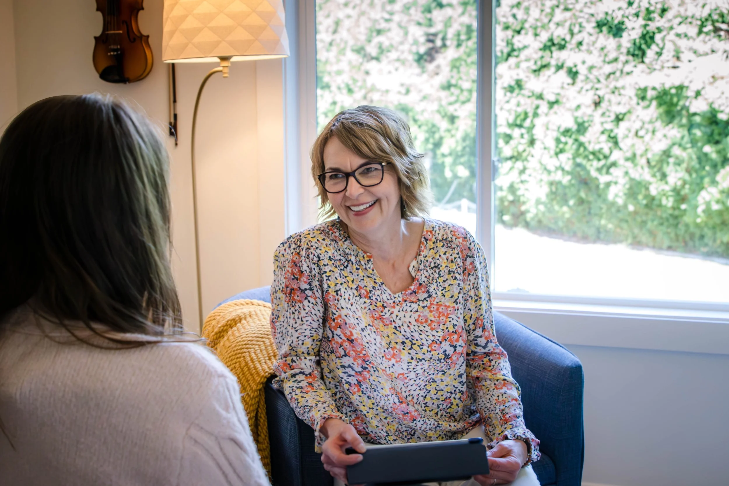 Two women having a conversation in a cozy room with a large window showing greenery outside, one woman wearing glasses and a floral top, the other with long hair and a beige sweater.
