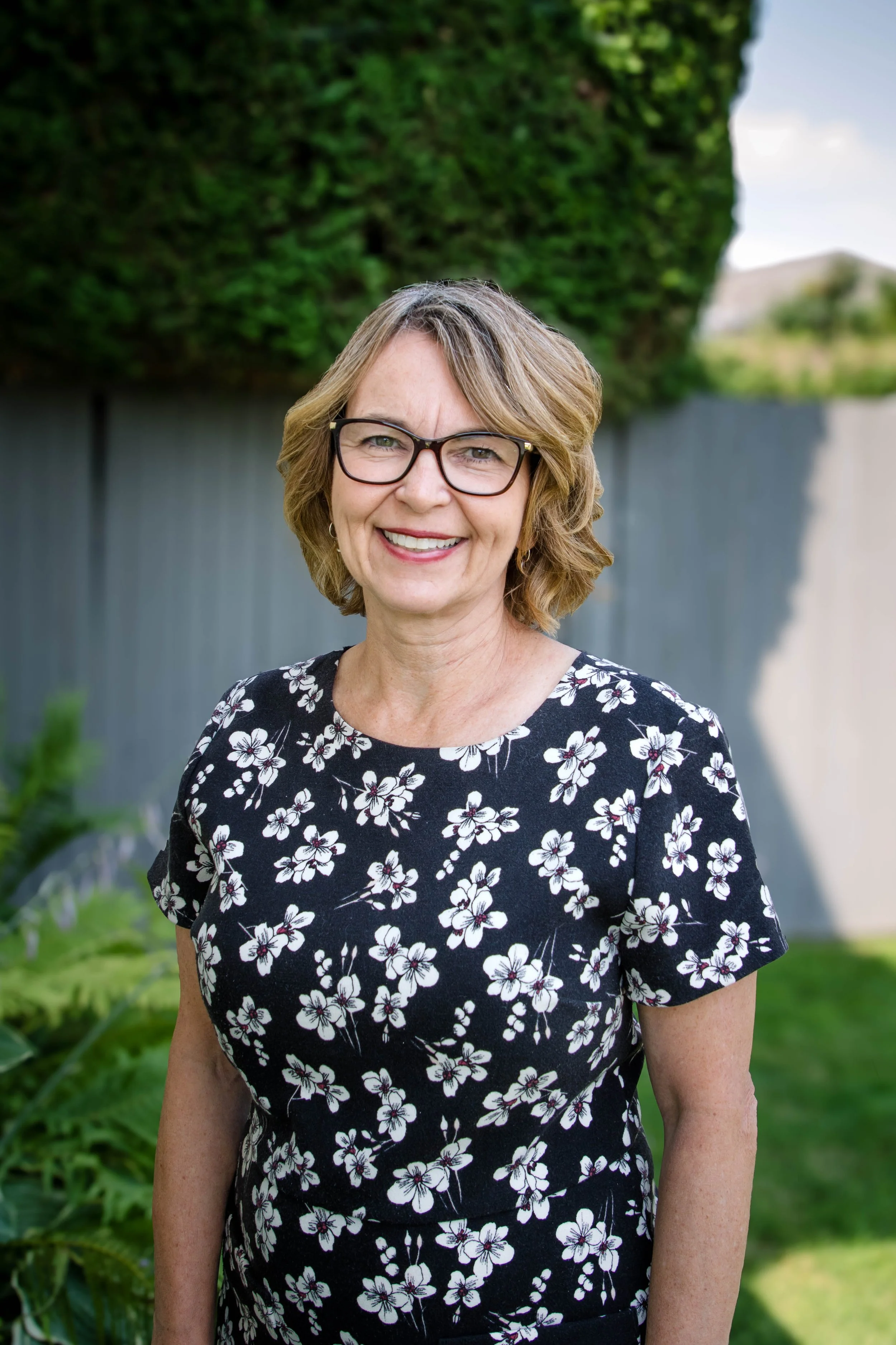 A smiling woman with short, wavy blonde hair and glasses, wearing a black dress with a white floral pattern, standing outdoors in front of a green hedge and a gray fence.