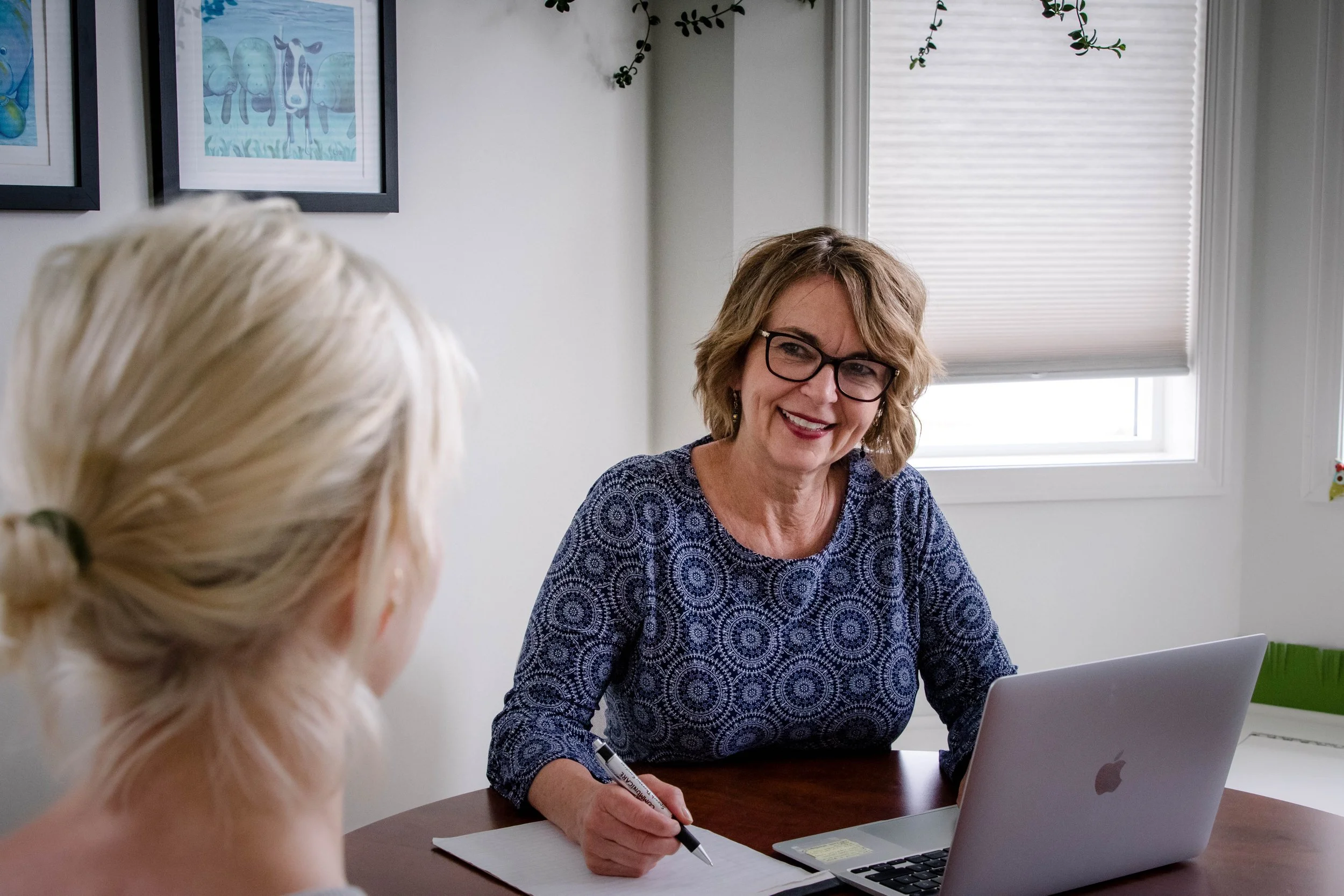 A woman with glasses and shoulder-length hair smiling, sitting at a desk with a laptop and notepad, talking with a person whose back is towards the camera, in a bright room with framed artwork on the wall.