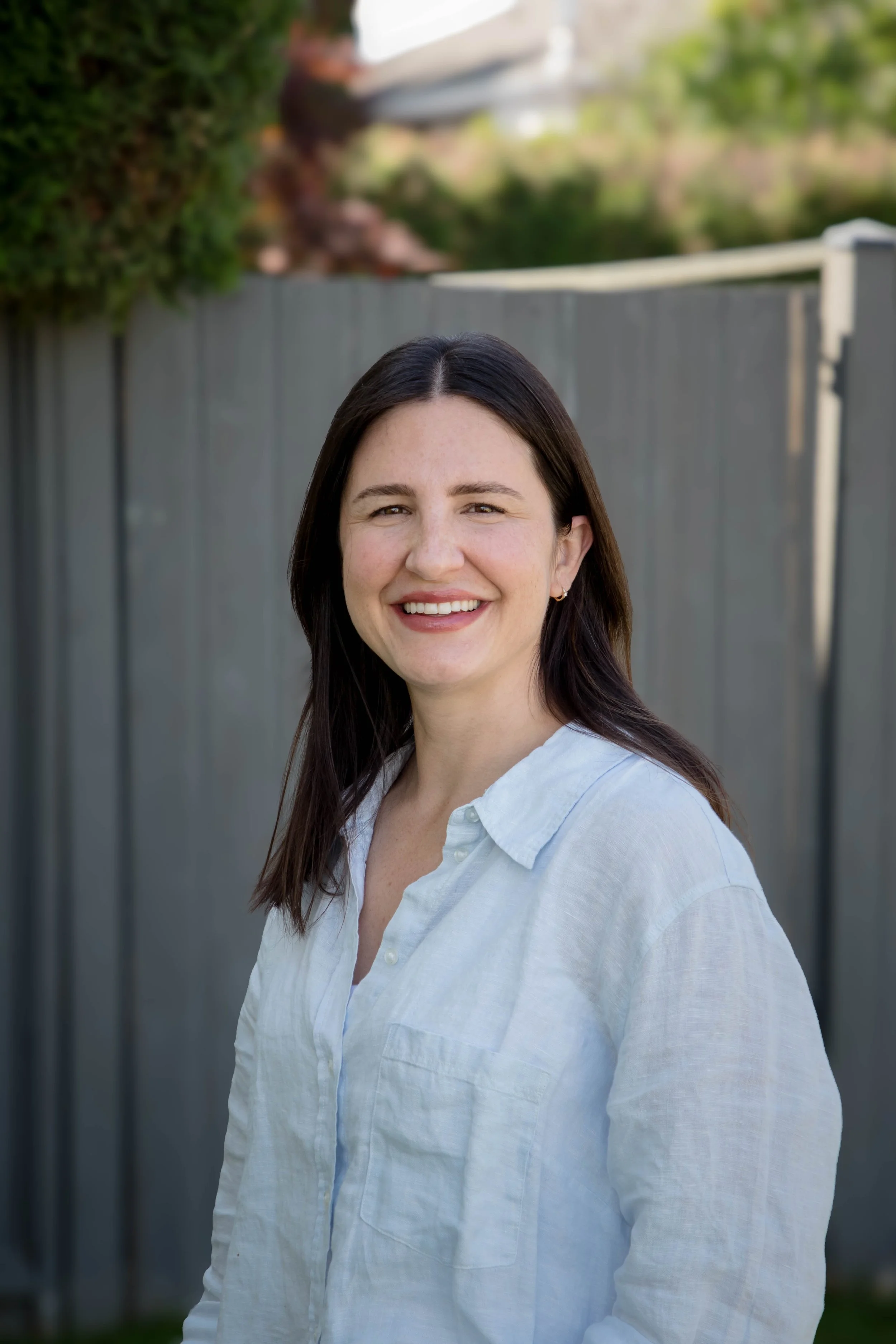 A woman with long dark hair smiling outdoors, wearing a white button-up shirt, in front of a gray wooden fence with trees in the background.