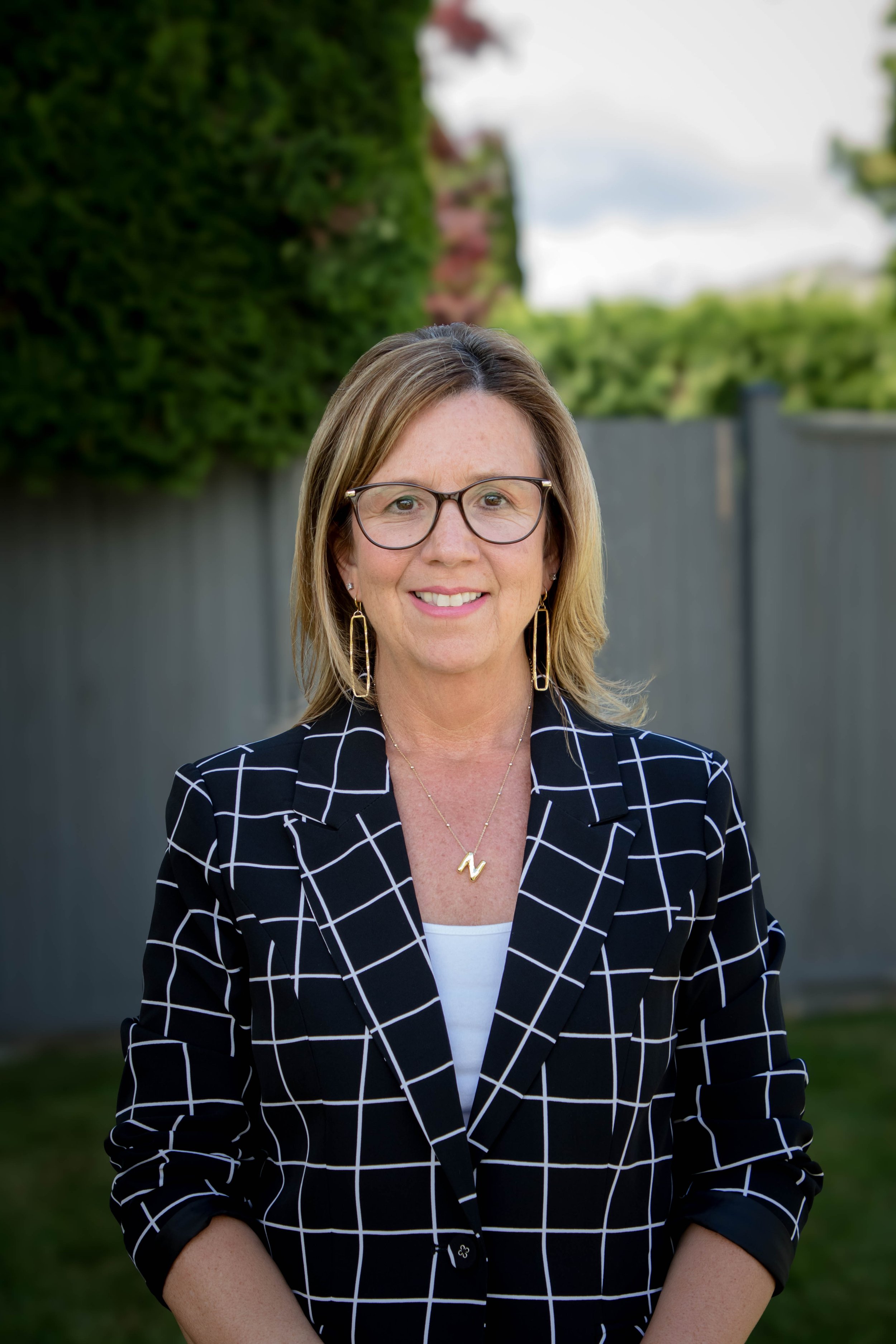 A woman with shoulder-length blonde hair, wearing glasses and gold jewelry, standing outdoors in front of a fence and greenery, smiling at the camera, dressed in a black blazer with a white checkered pattern over a white top.