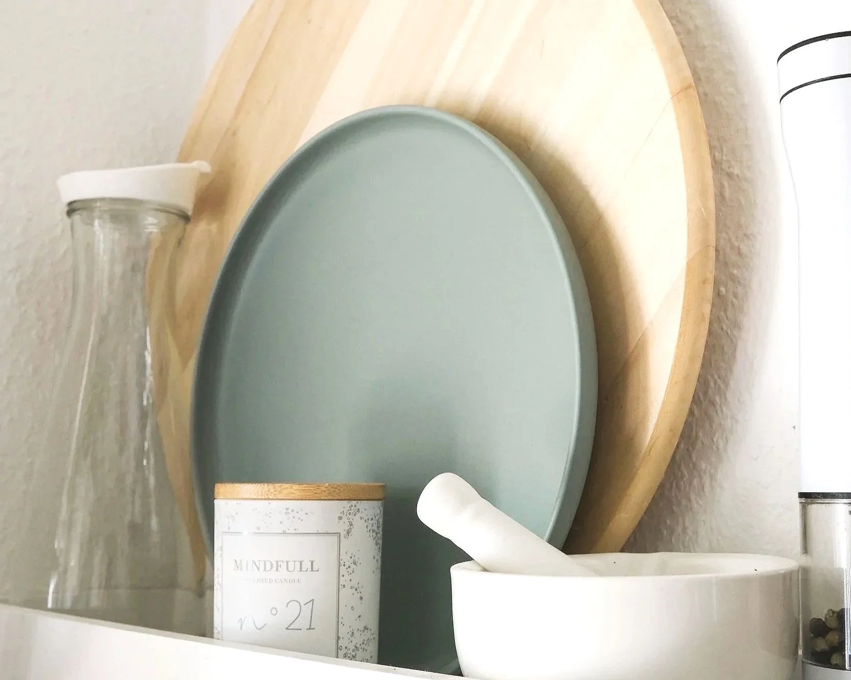 Decorative shelf with a glass bottle, a candle labeled 'MINDFULL No 21', a white mortar and pestle, a blue tray, and a white bowl, against a textured wall.