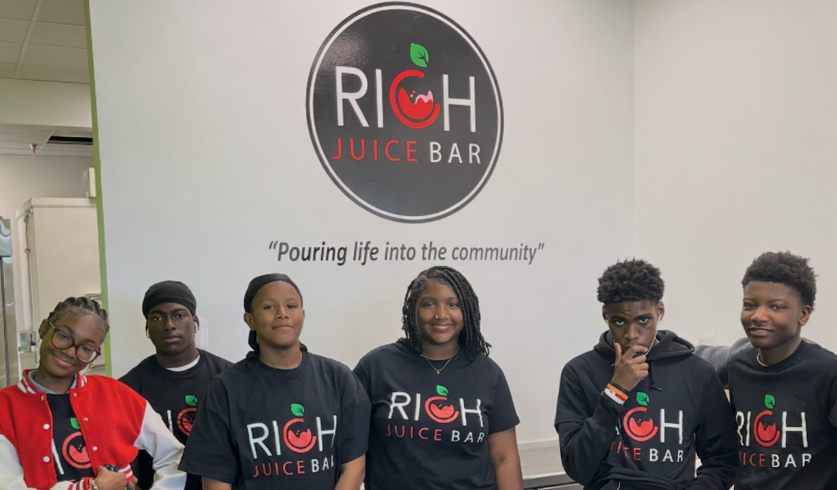 Six young people standing inside a juice bar, with a sign behind them reading 'Rich Juice Bar' and the tagline 'Pouring life into the community'.