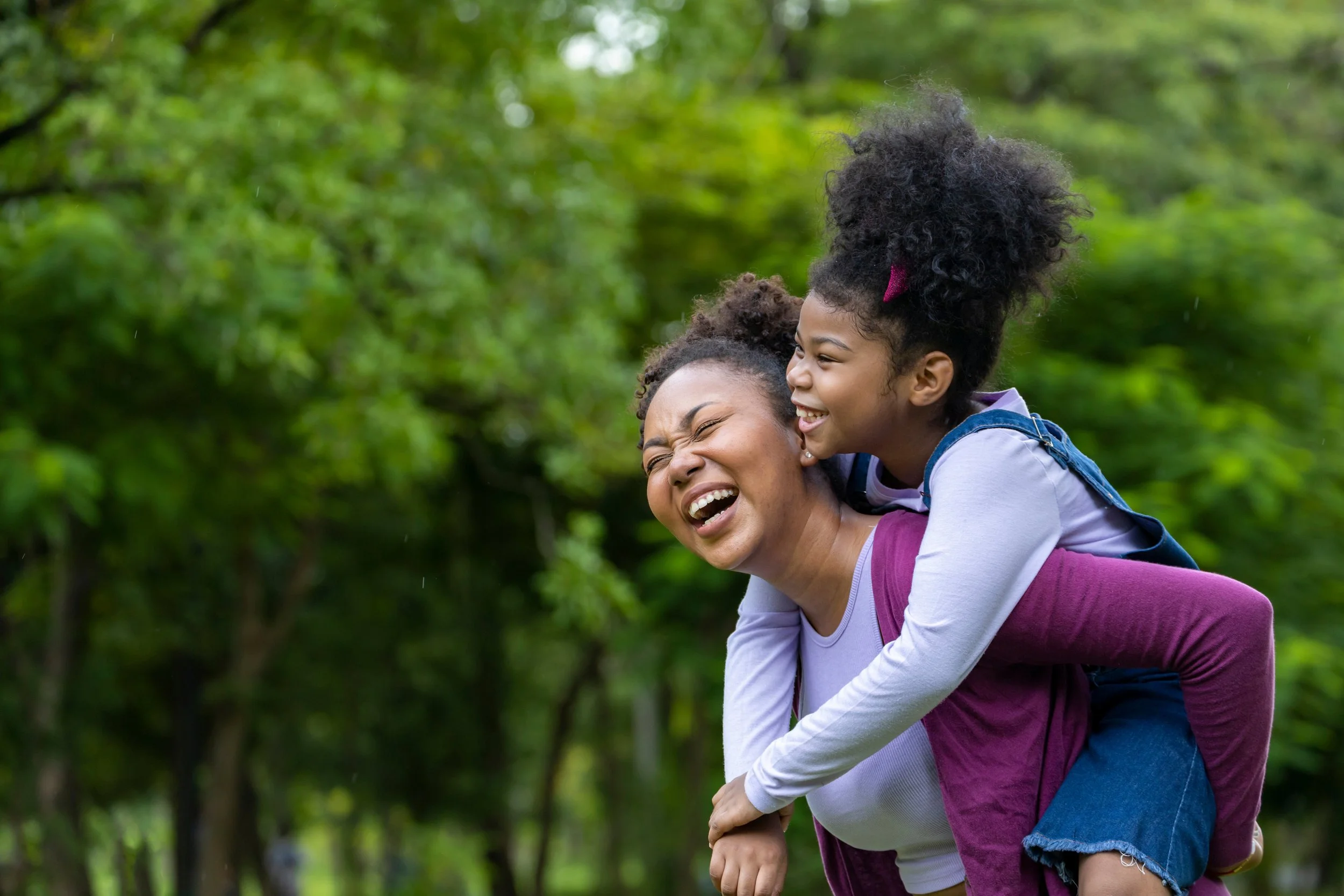 A woman giving a piggyback ride to a girl in a park with green trees in the background. Both are smiling and appear to be enjoying the moment.