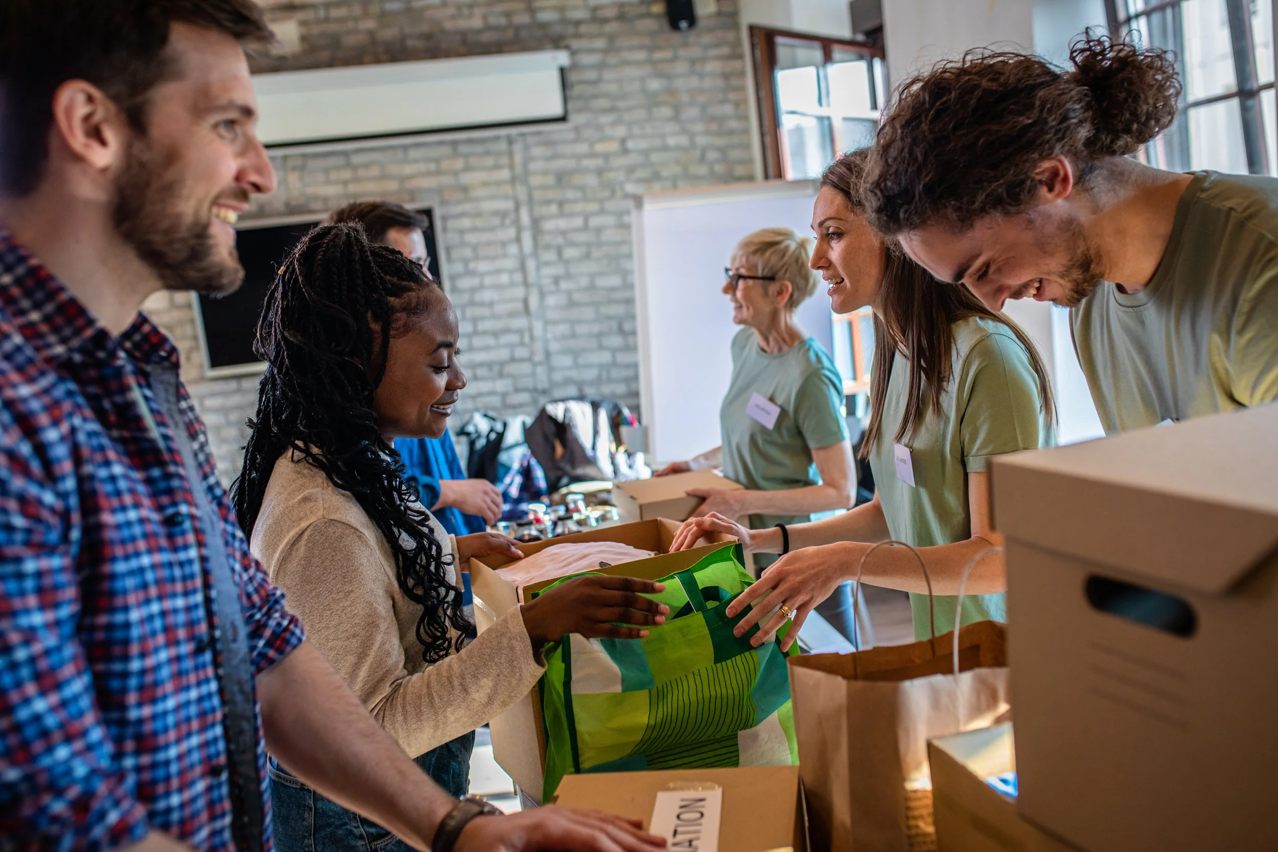 People at a charity or donation event, distributing or receiving items with boxes and bags in a bright room with large windows and brick walls.