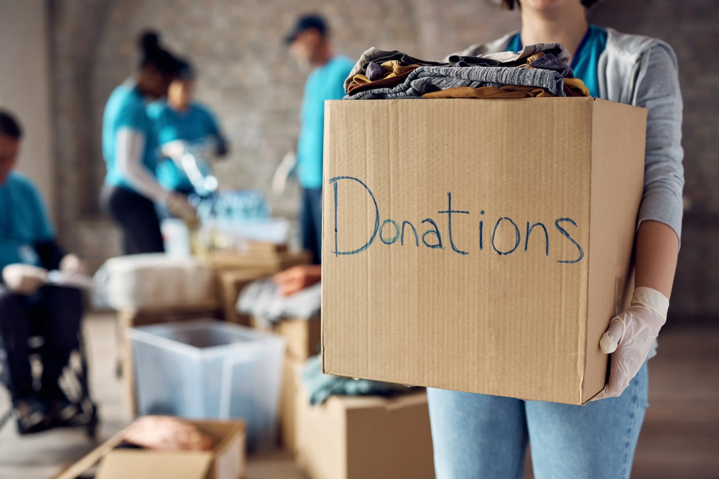 Person holding a cardboard box labeled 'Donations' with clothes inside, while other volunteers work in the background at a donation center.
