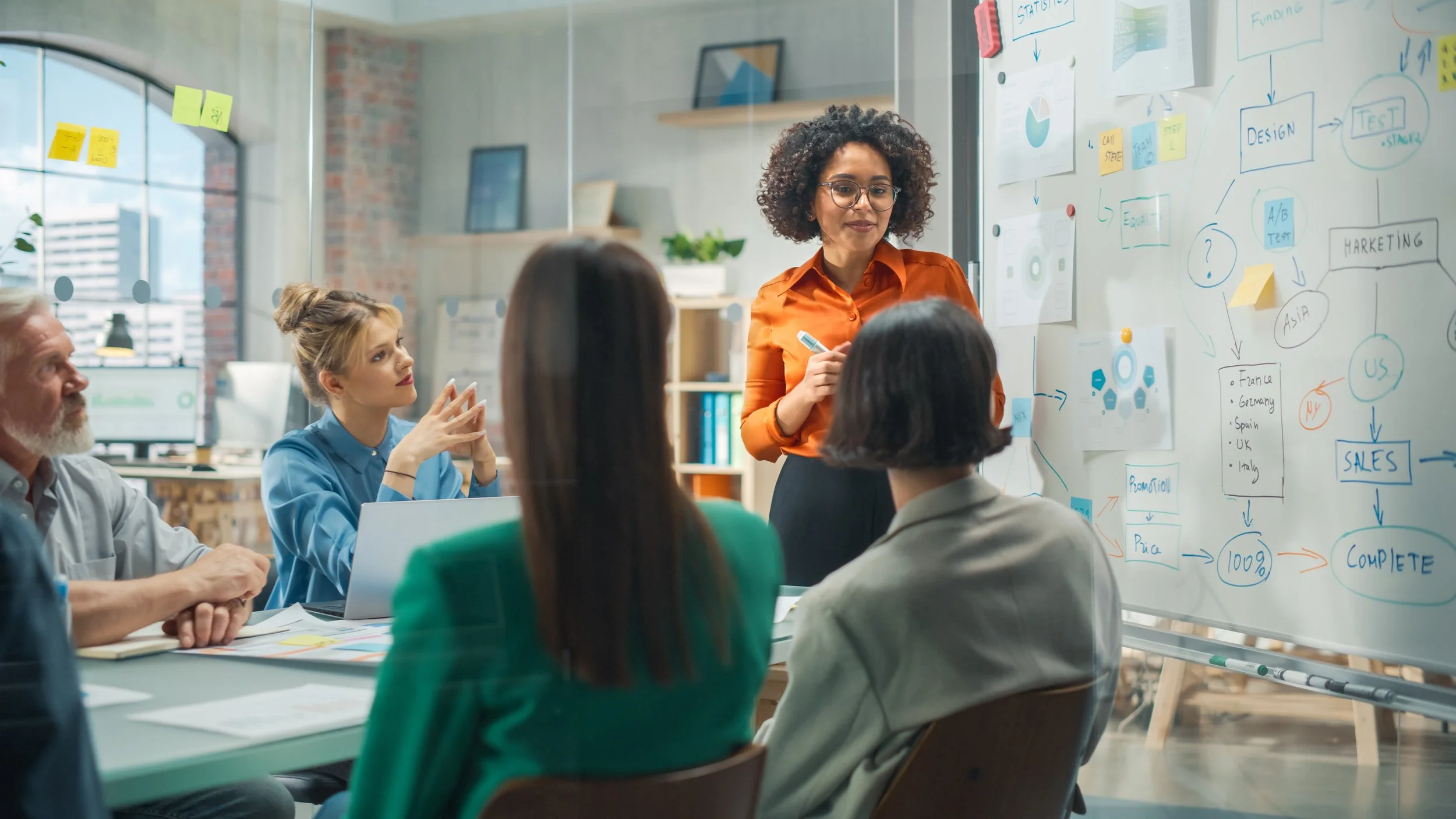 A diverse group of people in a business meeting room, listening to a woman with curly hair and glasses who is explaining a flowchart on a whiteboard.