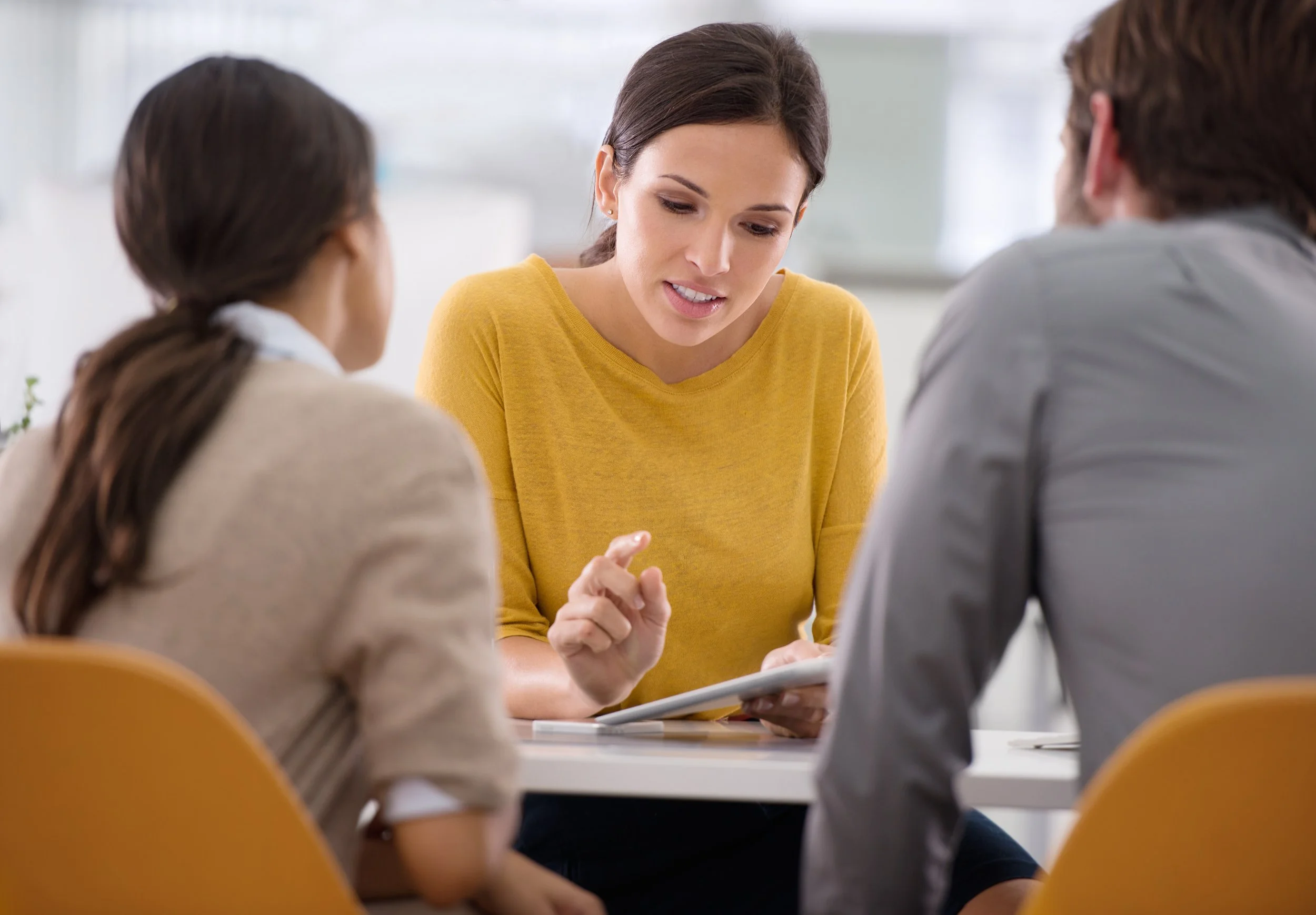 A woman in a yellow shirt speaking to two people with brown hair, in a meeting or discussion, seen from behind.