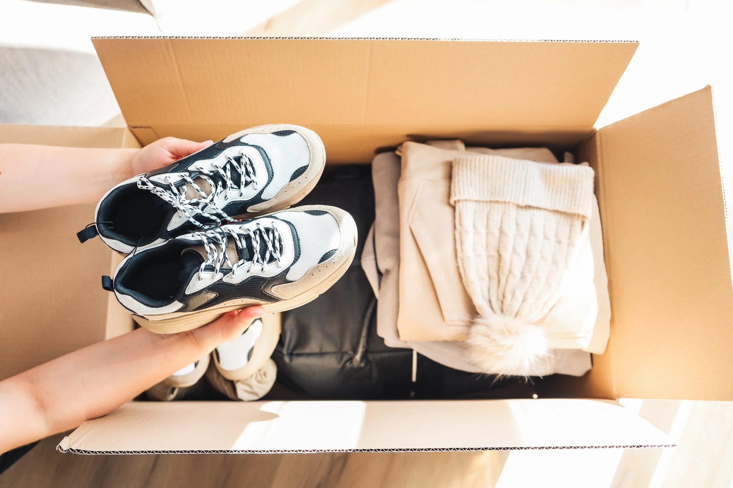 Person holding a pair of white and black sneakers inside a cardboard box with folded clothing and a blanket.