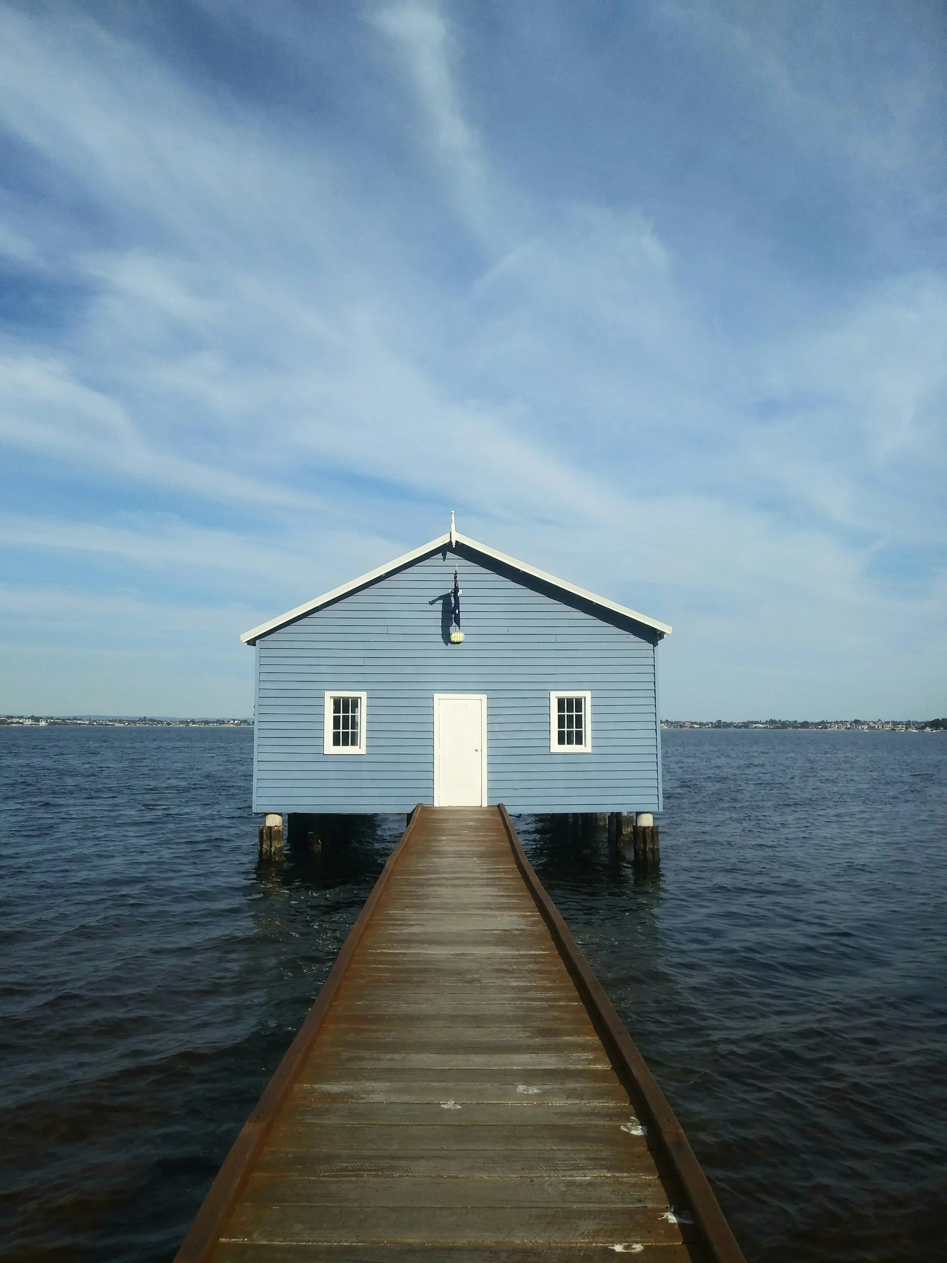 A blue house with white trim sits on water. There is a wooden dock leading to the house. The sky above is partly cloudy.