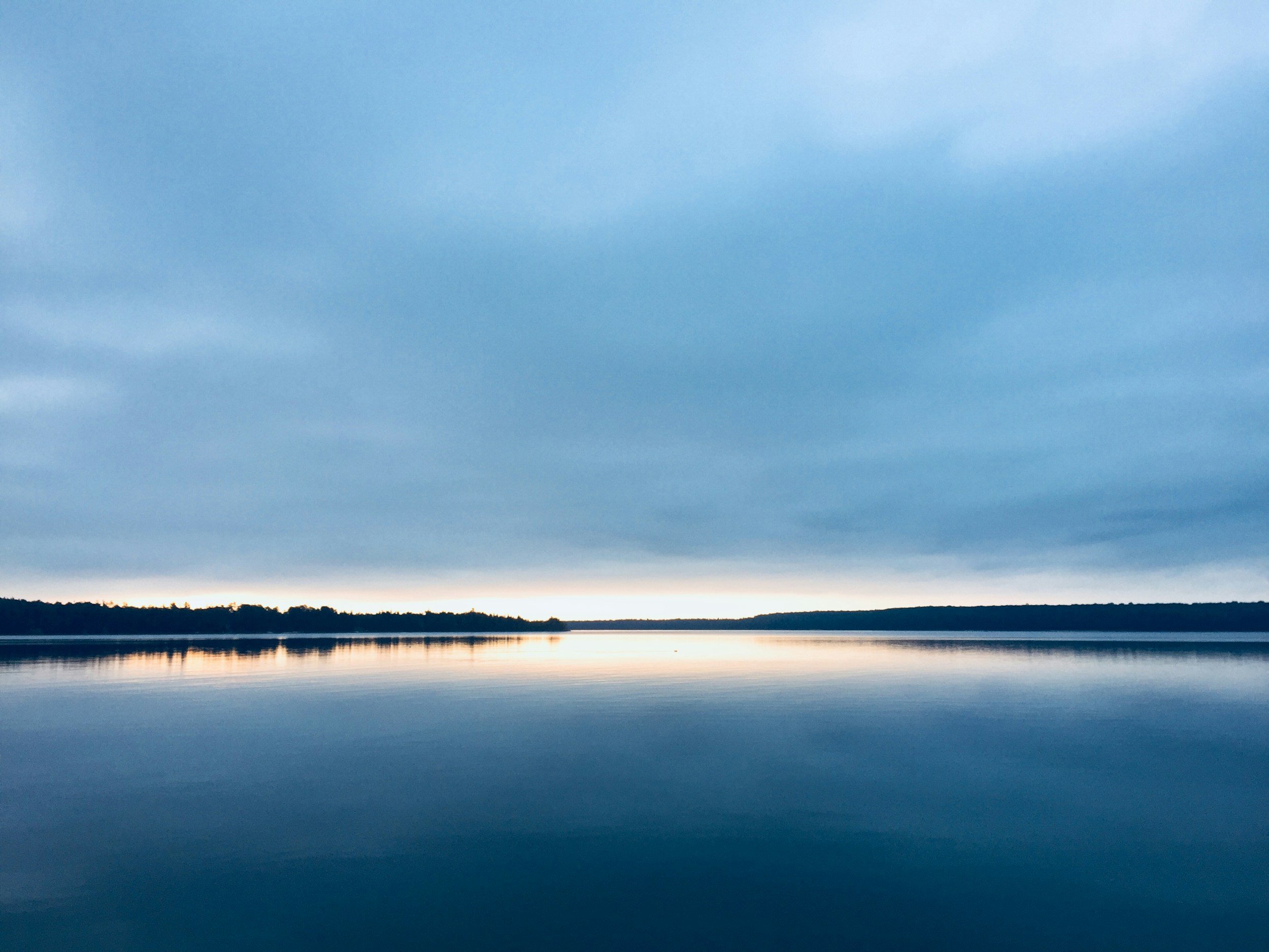 A peaceful lakeside scene during the evening, with a calm body of water reflecting the darkening sky and a silhouette of trees along the horizon.