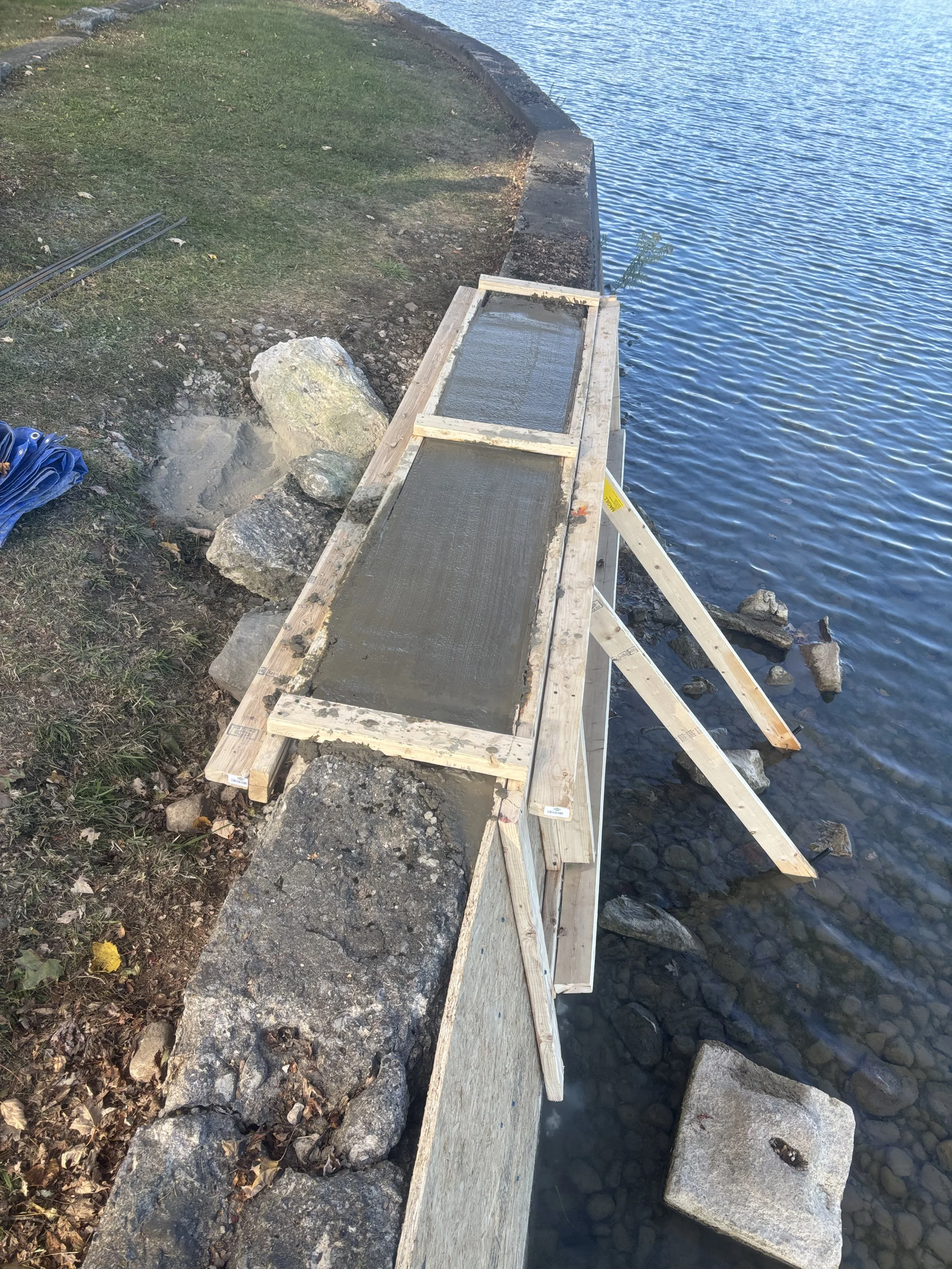 Construction of a small concrete pathway along a lakeshore, with a wooden frame in place, surrounded by rocks and grass.