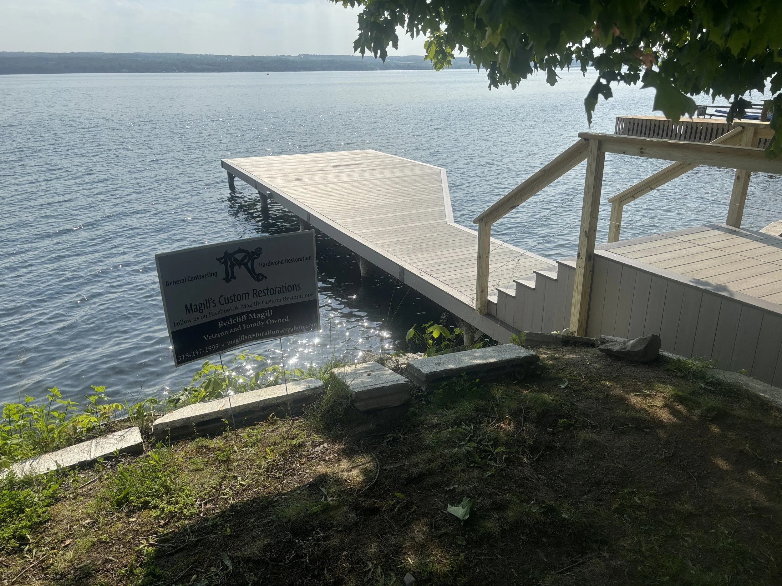A wooden dock extending over a body of water with trees and hills in the background. Part of a sign is visible in the lower left corner, advertising a restoration business.