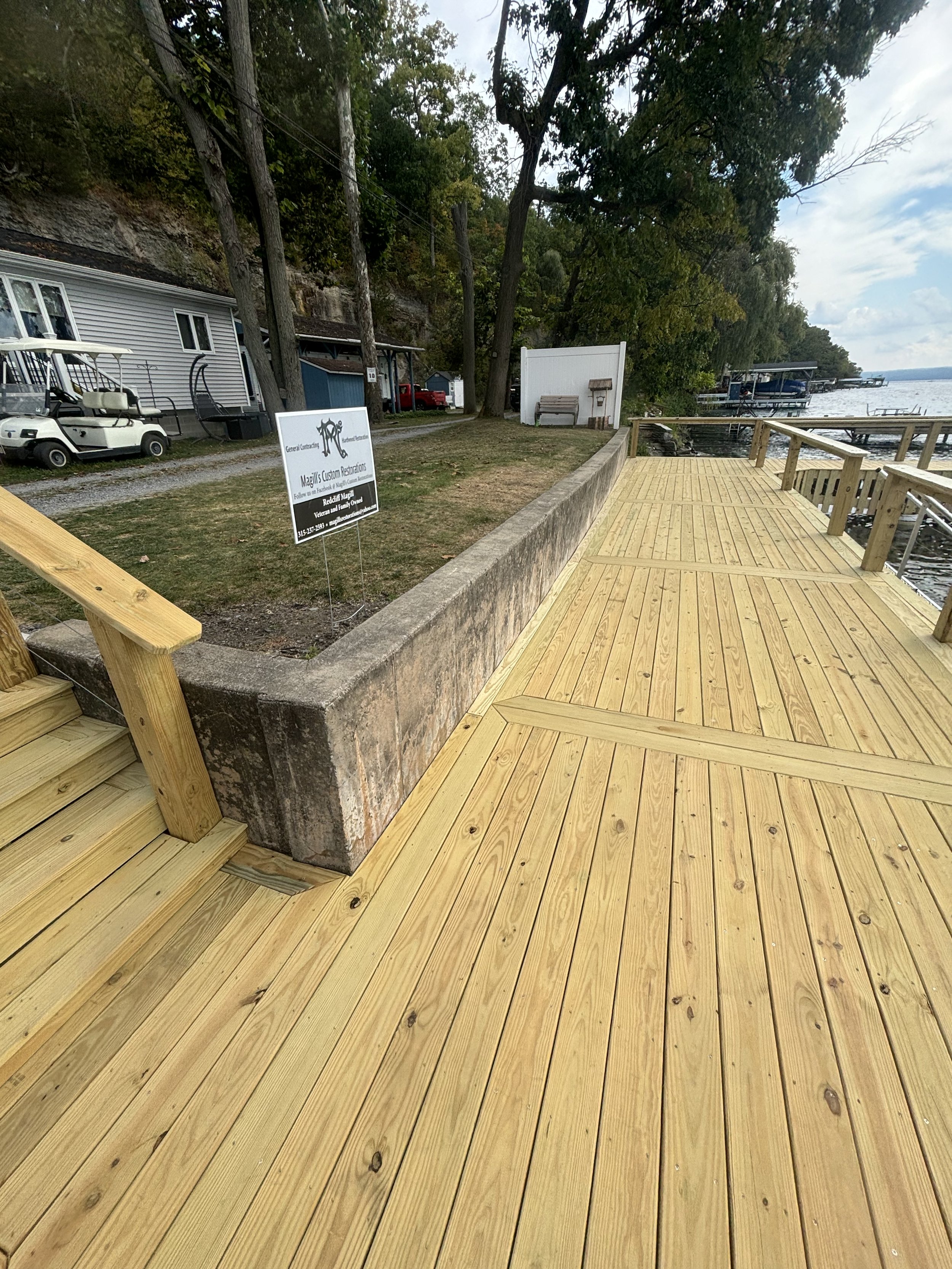 View of a wooden dock along a waterfront with trees in the background, a white sign, a white fence, and some boats and structures near the water.