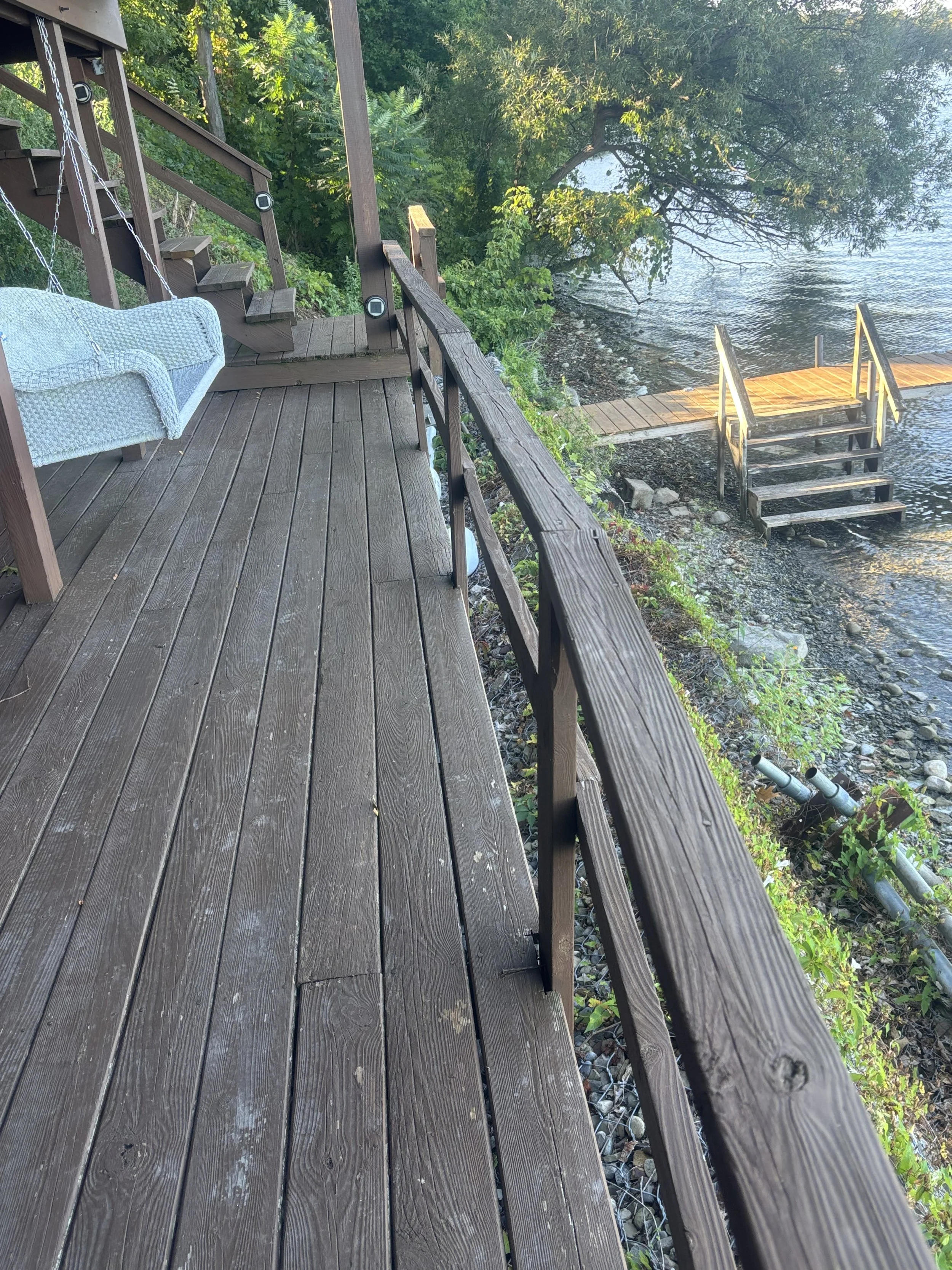 Wooden deck by a lake with stairs and a swing seat, surrounded by green trees and water.