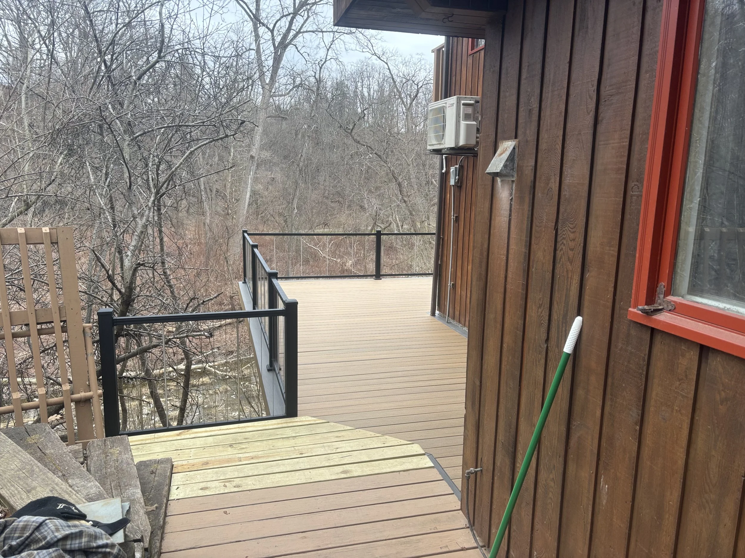 Balcony or deck area attached to a wooden house, showing railings, a staircase leading down, and trees with snow in the background.