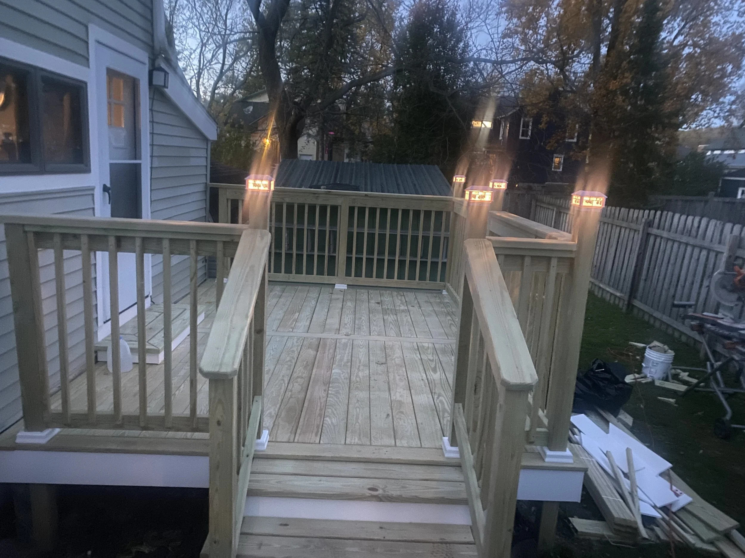 A wooden deck with a railing under construction, houses, trees, and a fence are visible in the background, with evening light.