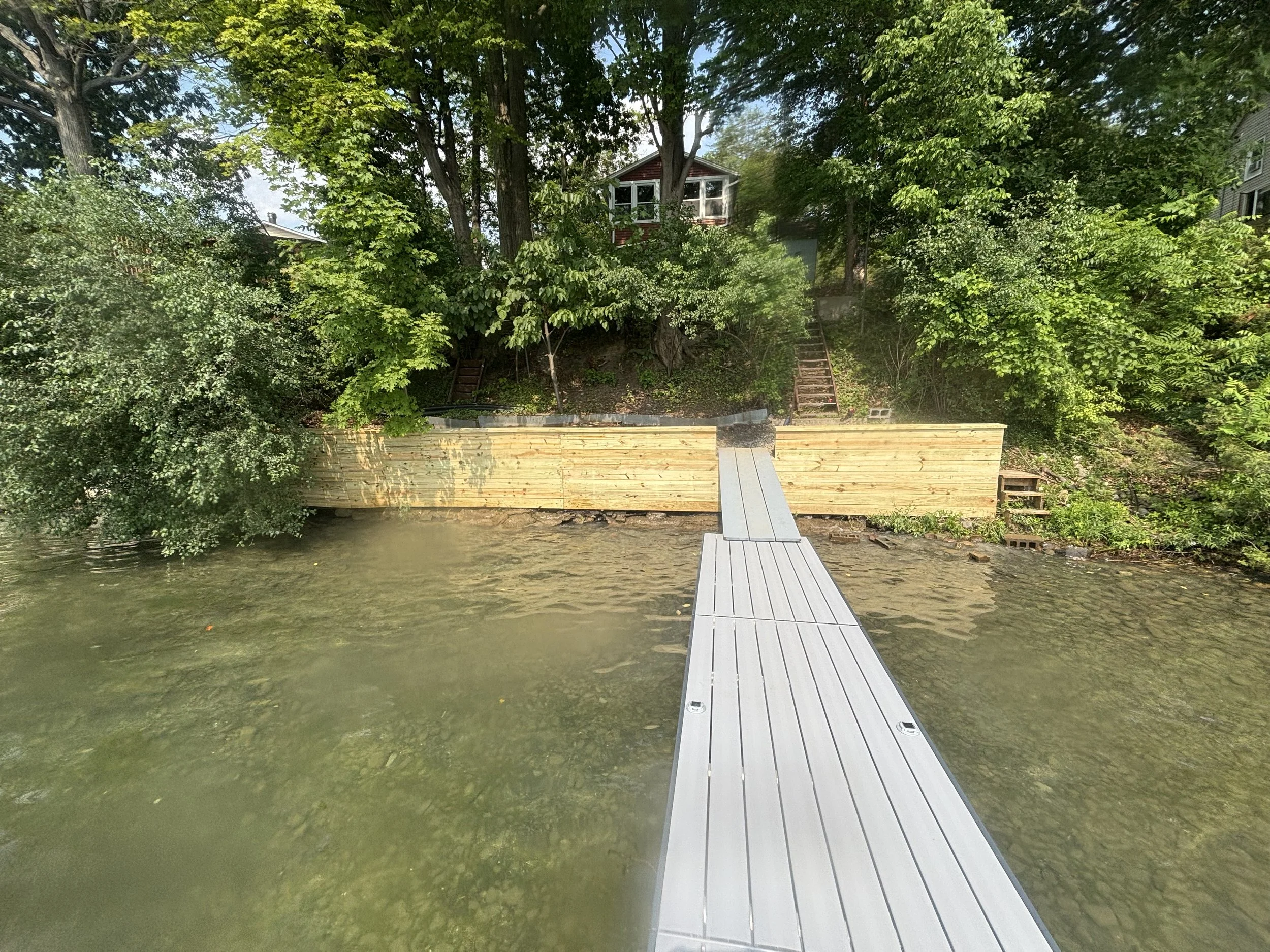 A floating dock extends over a body of water towards a wooded shoreline with a house and stairs leading up the hill.