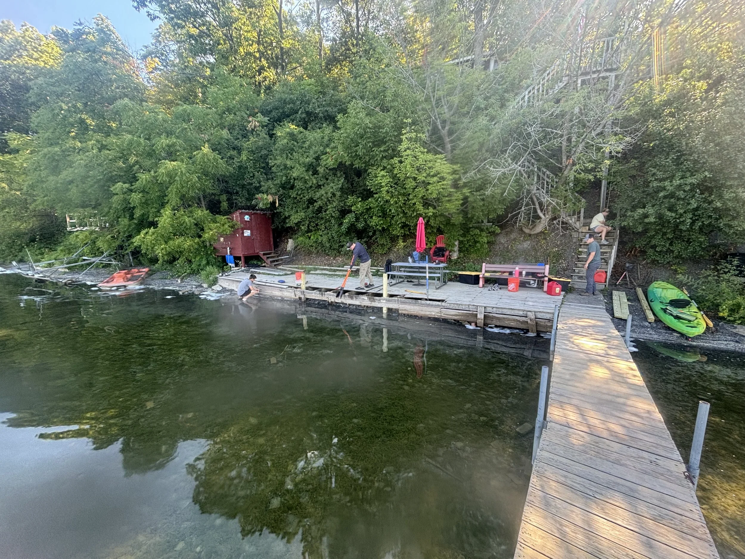 People on a dock by a river, some fishing, with trees and a house in the background.