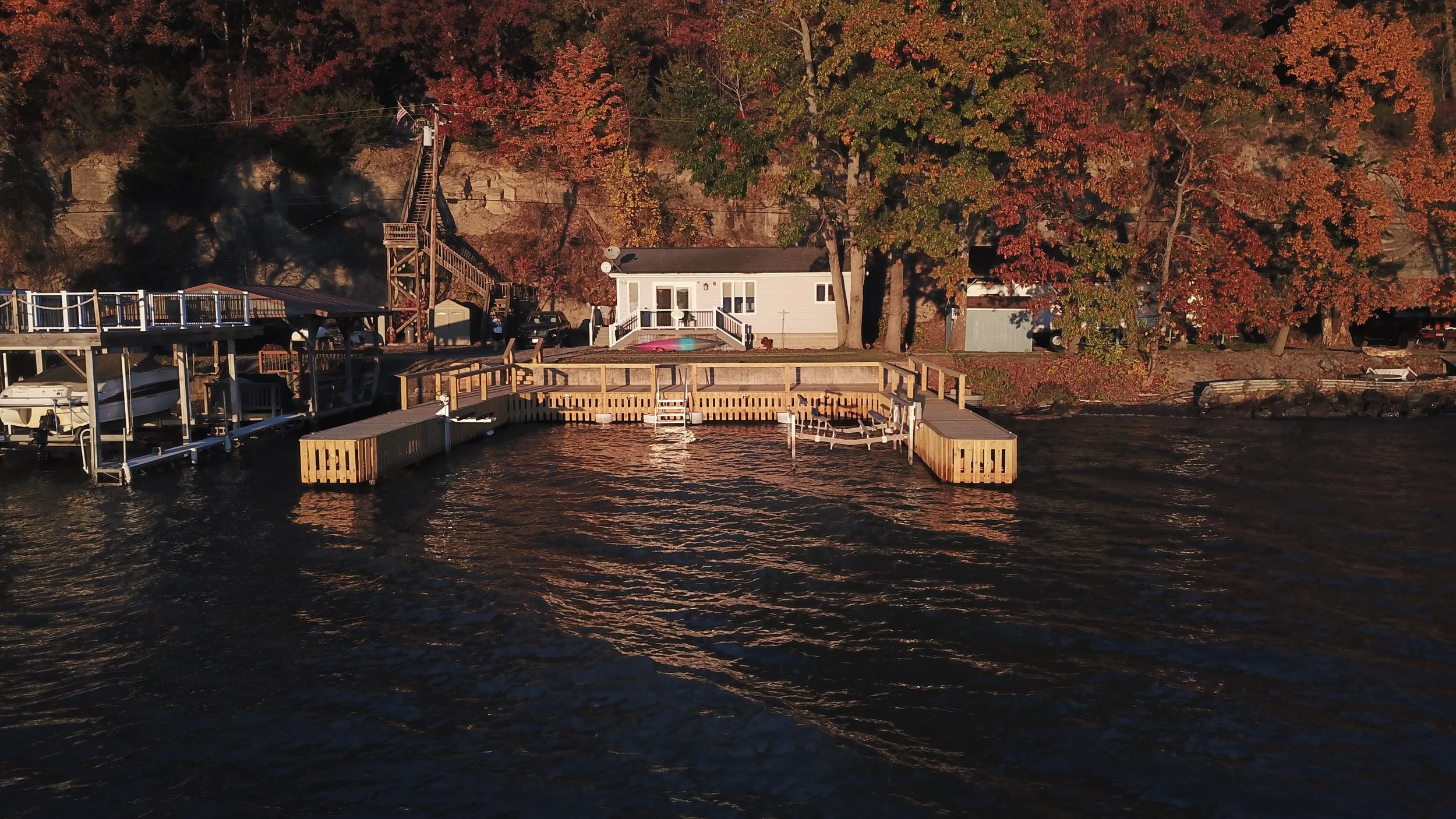 A lakeside scene during autumn with a dock, boats, and a house surrounded by colorful fall foliage.