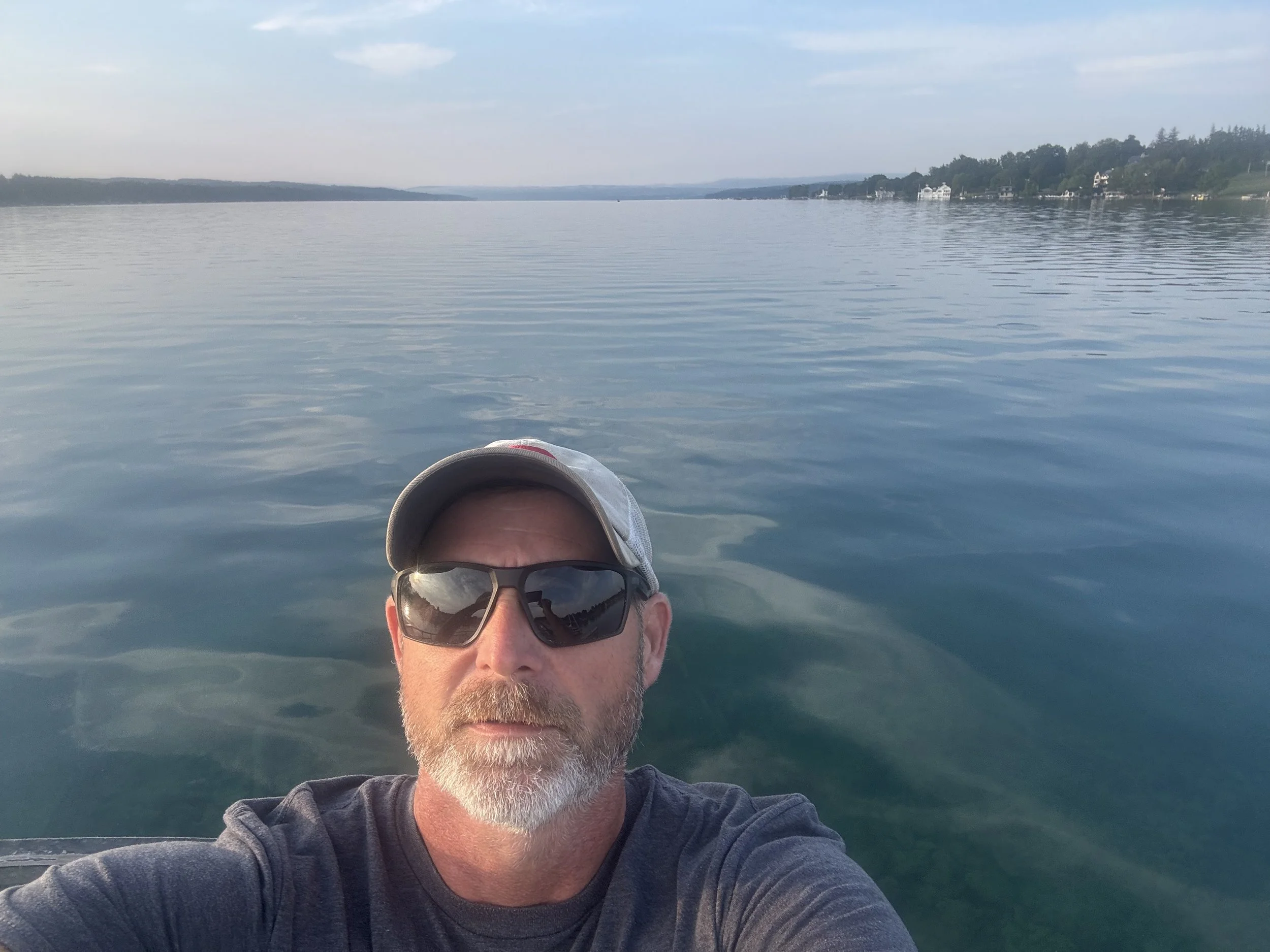 A man with sunglasses, a cap, and a beard taking a selfie on a boat or dock on a calm lake, with houses and trees on distant shores under a blue sky.