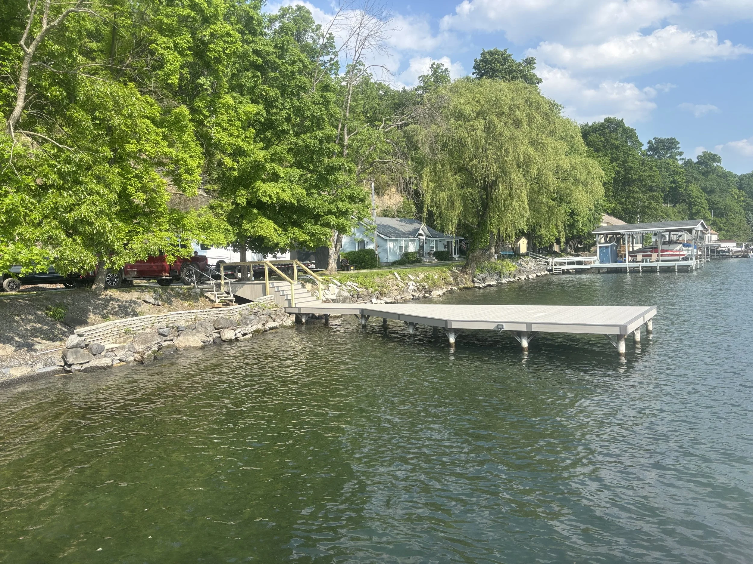 A lakeside scene with green trees, houses, a dock, and boats, under a partly cloudy blue sky.