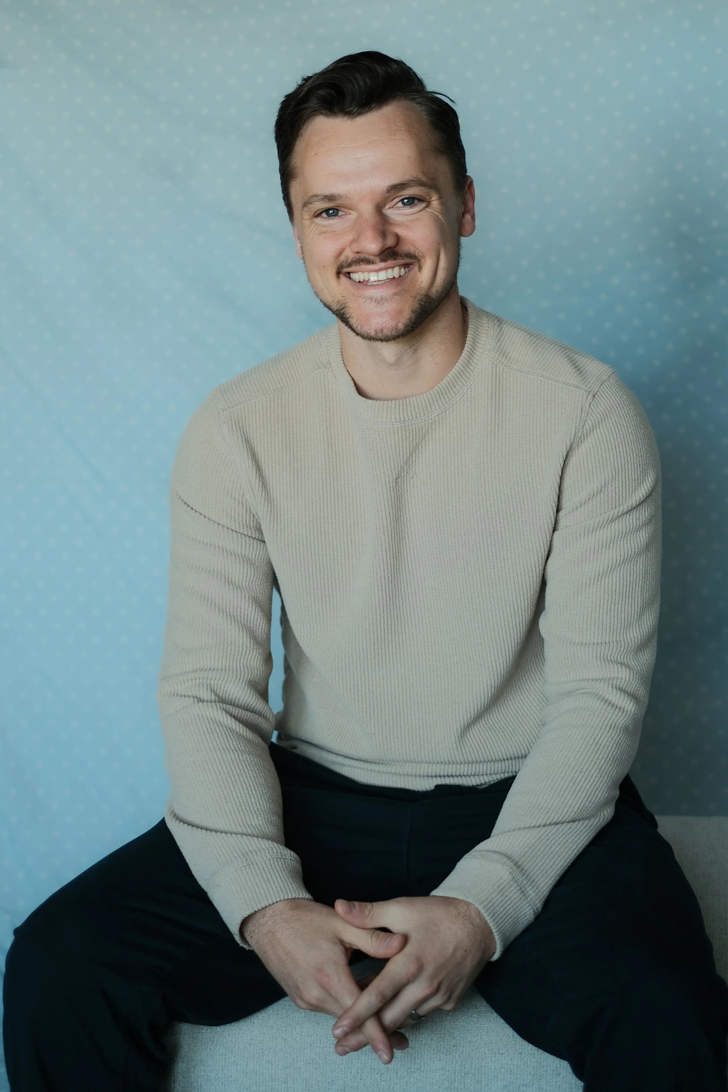 Man with dark hair and light skin smiling while sitting in front of a light blue polka dot background, wearing a beige sweater and dark pants.