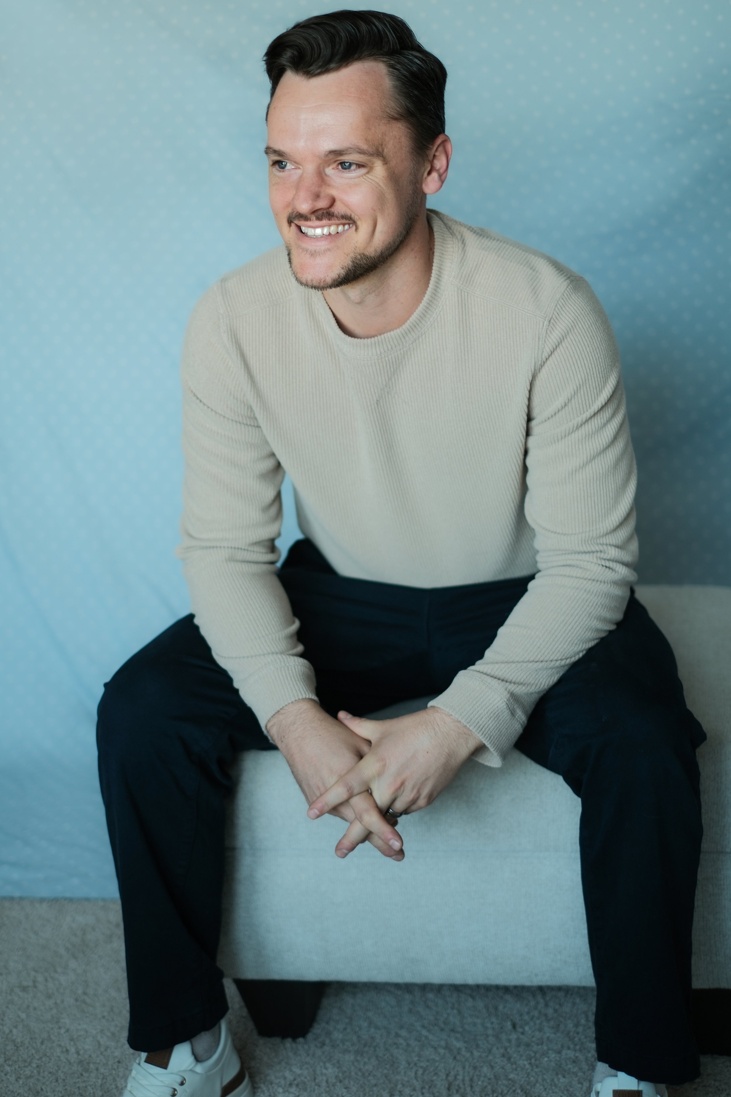 A young man with dark hair and a beard smiling, sitting on a stool against a light blue patterned background, wearing a beige sweater, black pants, and white sneakers.
